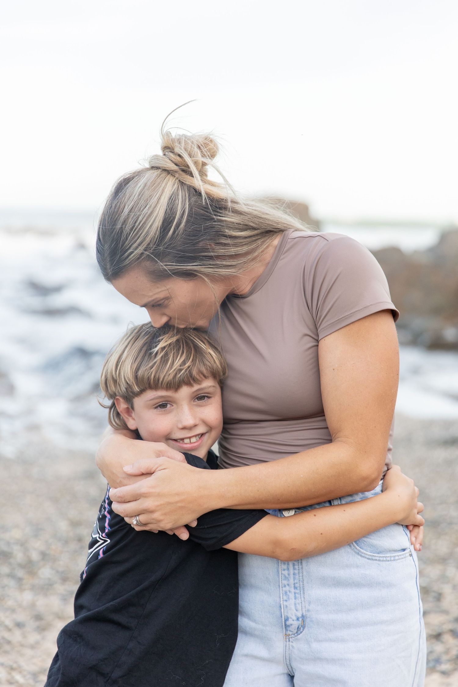 A mother lovingly embraces her smiling young son on a rocky beach, both enjoying a tender moment together. Coffs Coast photographer.