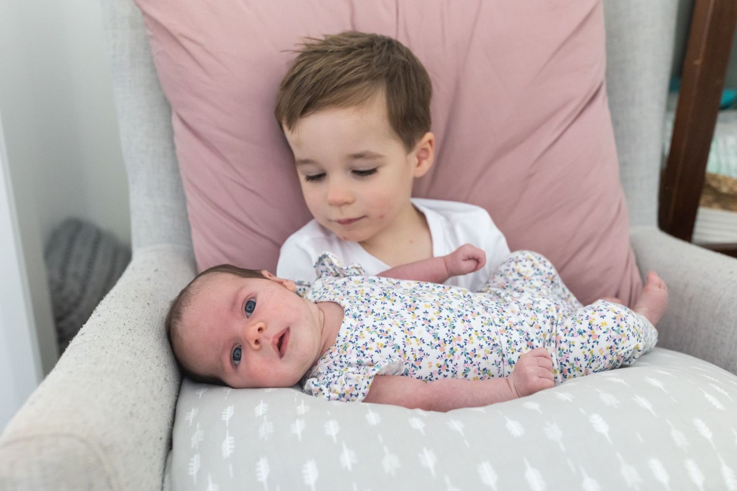 Young toddler boy sitting in a gray chair holding a newborn baby wrapped in a floral swaddle blanket.