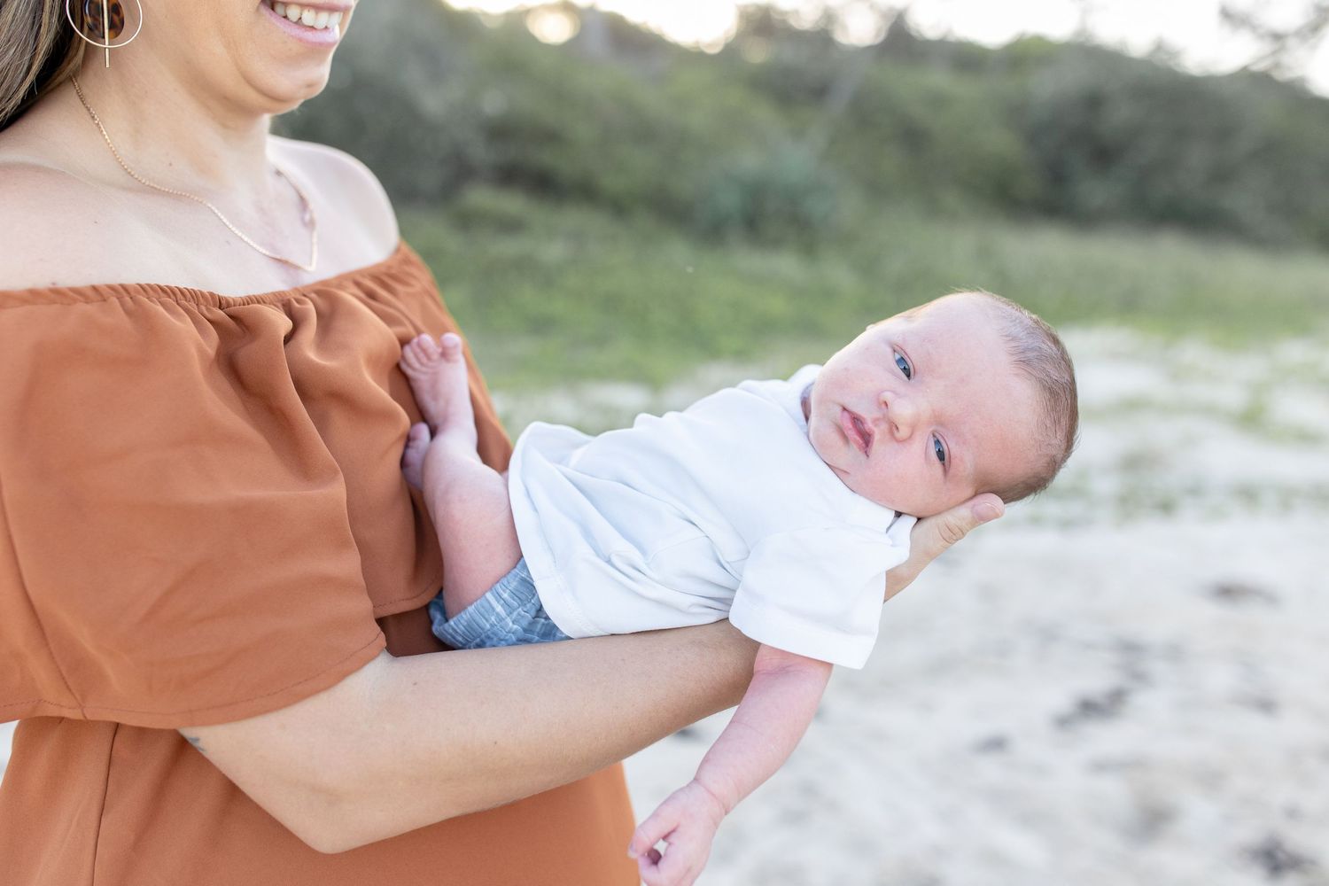 A smiling mother in an orange off-shoulder dress holds her newborn baby outdoors on a sandy beach. Coffs Coast photographer.