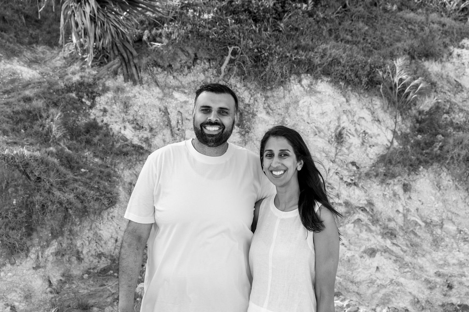 Black and white photo of a smiling couple posing together outdoors with trees and a rocky hillside behind them. Coffs Coast photographer.