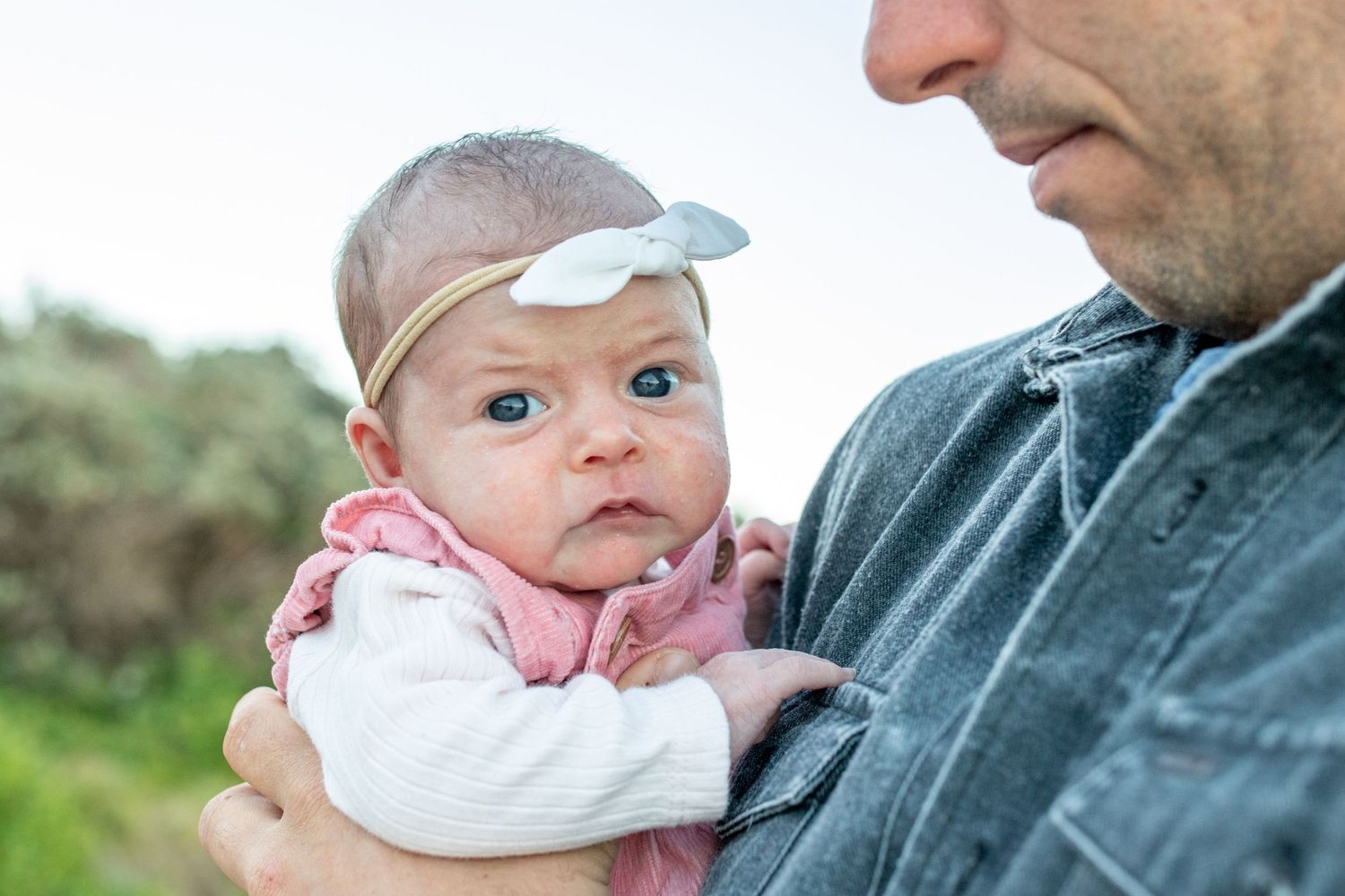 Father holding newborn baby girl wearing pink outfit and white bow headband outdoors. Coffs Coast photographer.