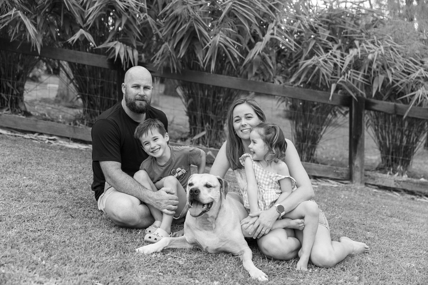 Black and white family photo with parents, two children, and a Labrador dog sitting on grass near bamboo fence.