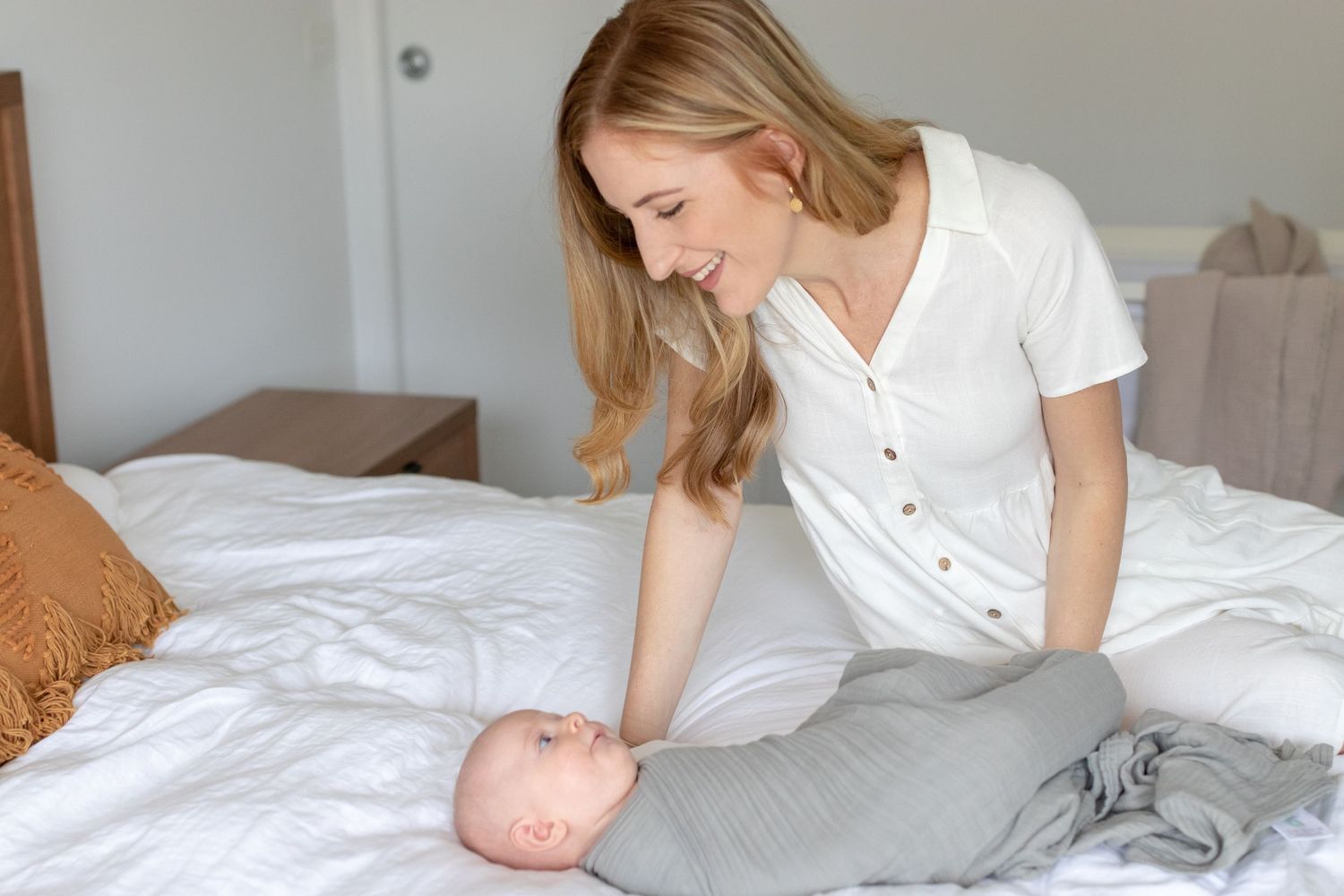 Smiling mother interacting with her newborn baby wrapped in a white swaddle on a bed.