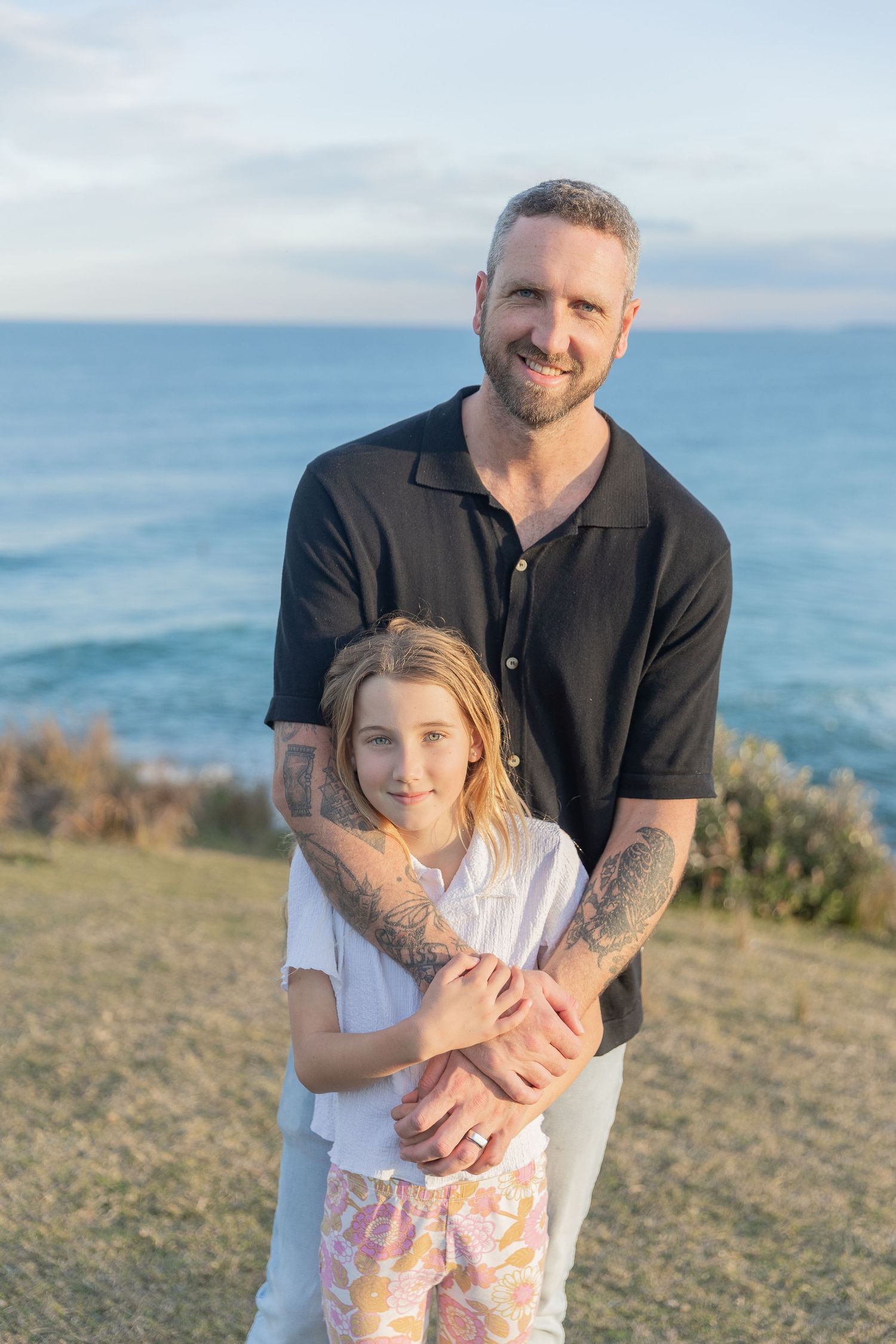 Parent and child embracing outdoors with ocean view and blue sky in background.