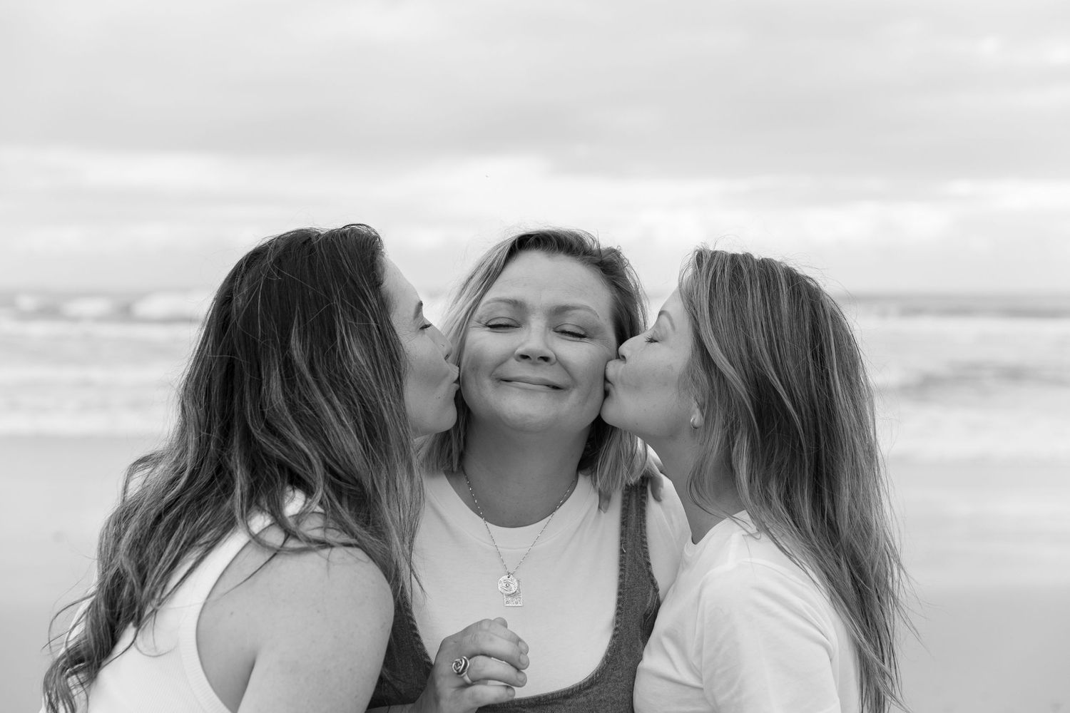 Two women kissing a smiling woman on the cheeks at the beach in a black and white photo.