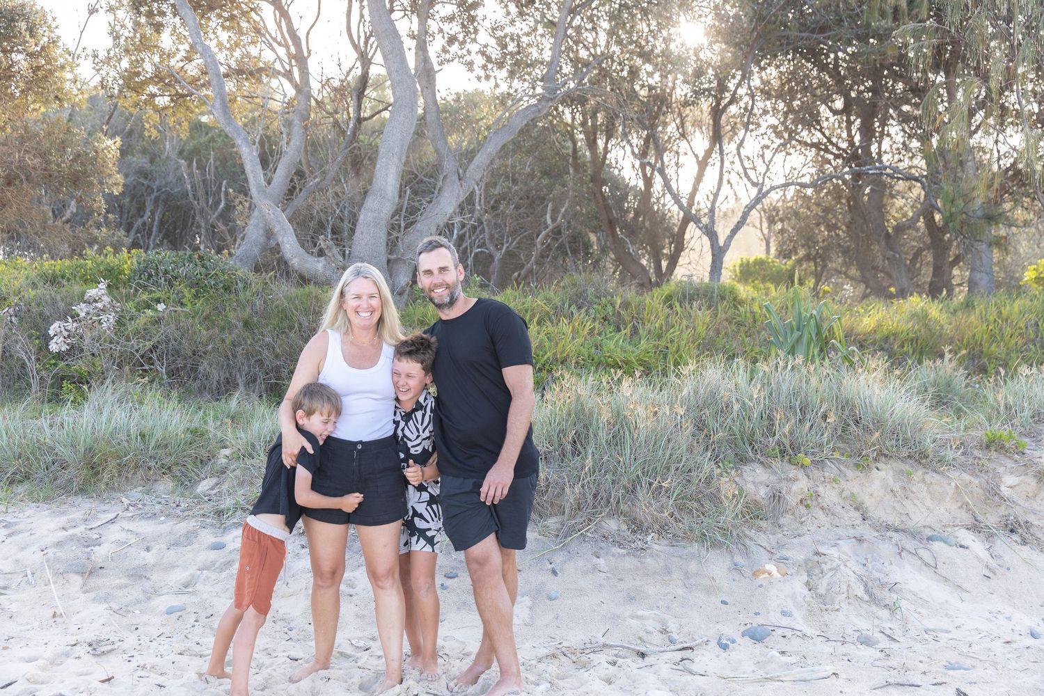 Happy family of four smiling together on a sandy beach surrounded by lush green trees at sunset.