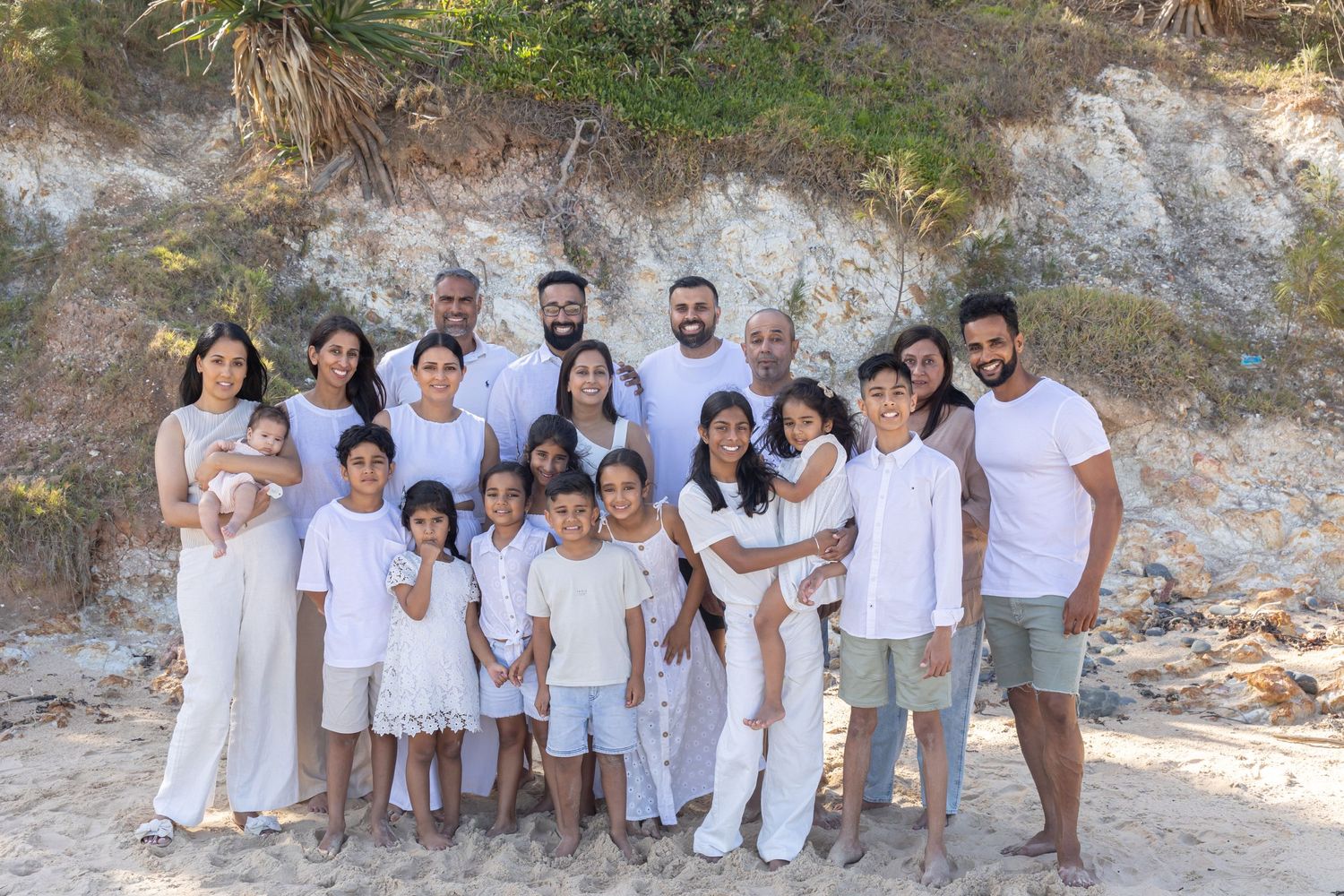 Large family group photo on sandy beach, all wearing white outfits, smiling together with rocky cliffs behind them. Coffs Coast photographer
