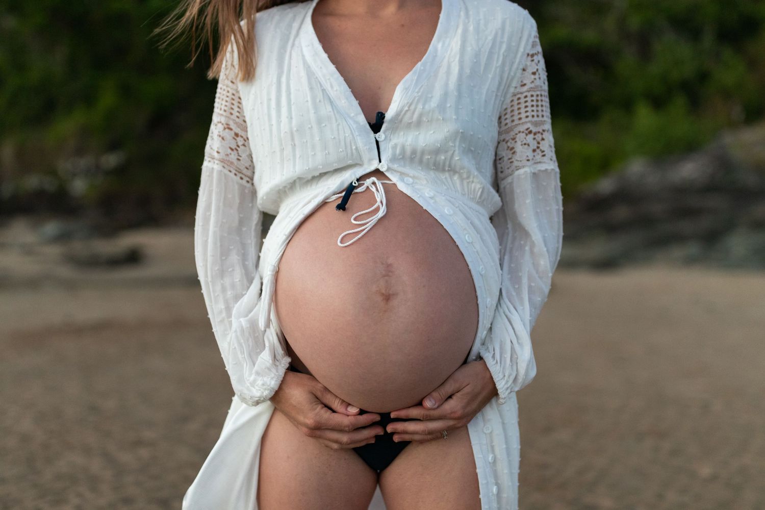 Pregnant woman in white lace dress holding baby bump on beach during maternity photoshoot.