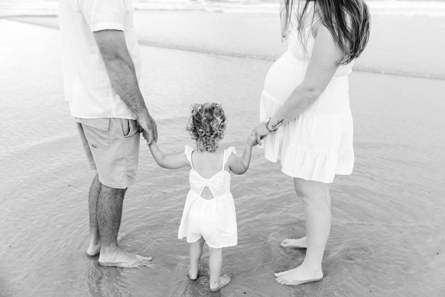 Black and white photo of pregnant couple holding hands with toddler girl on beach. Coffs Coast photographer.