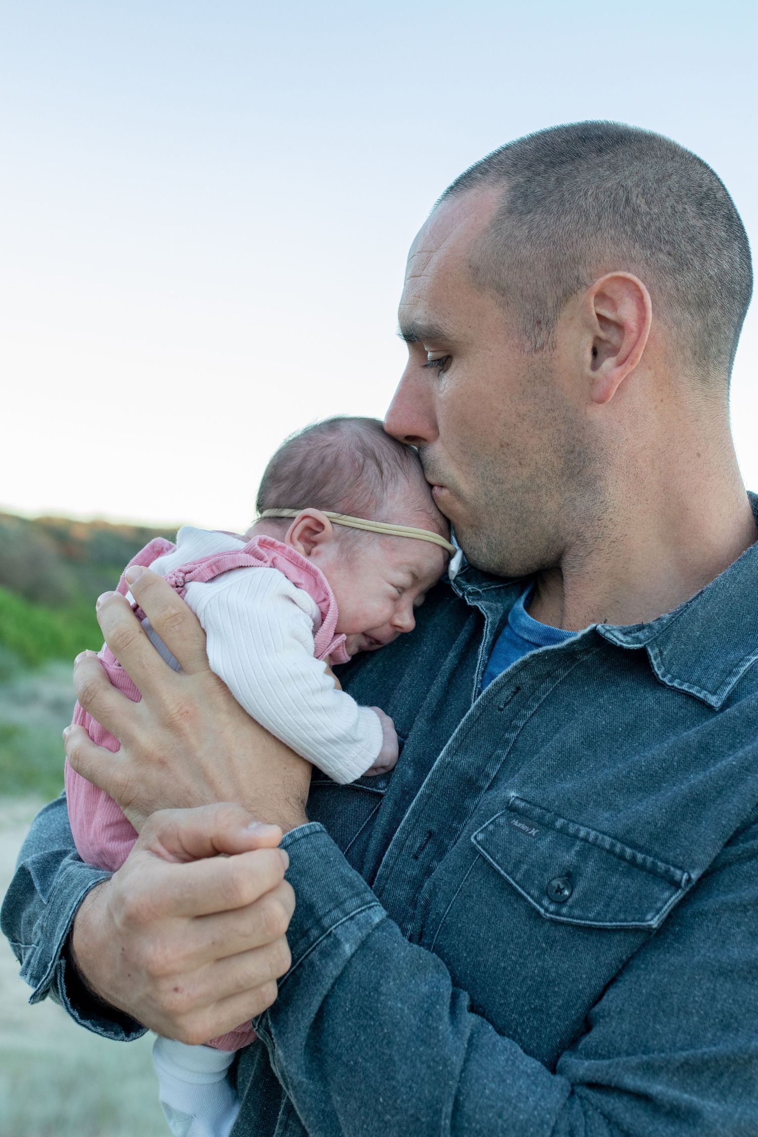 A father tenderly kisses his newborn baby girl while holding her gently outdoors near a beach. Coffs Coast photographer.