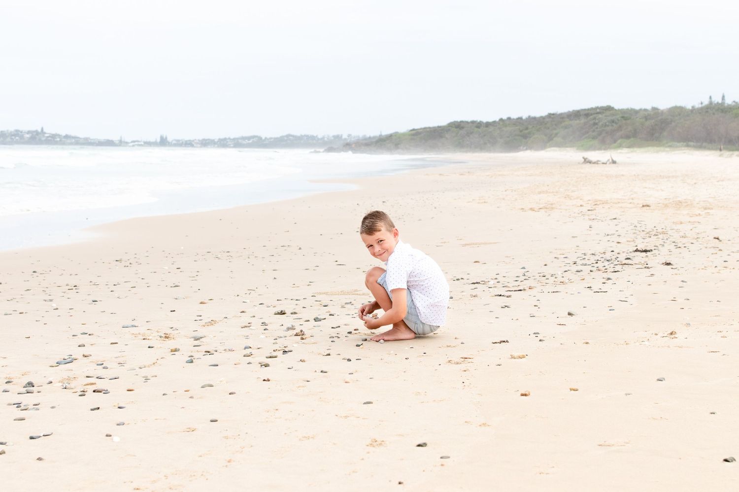 Young boy sitting on sandy beach shoreline, smiling and playing with shells near ocean waves. Coffs Coast photographer.