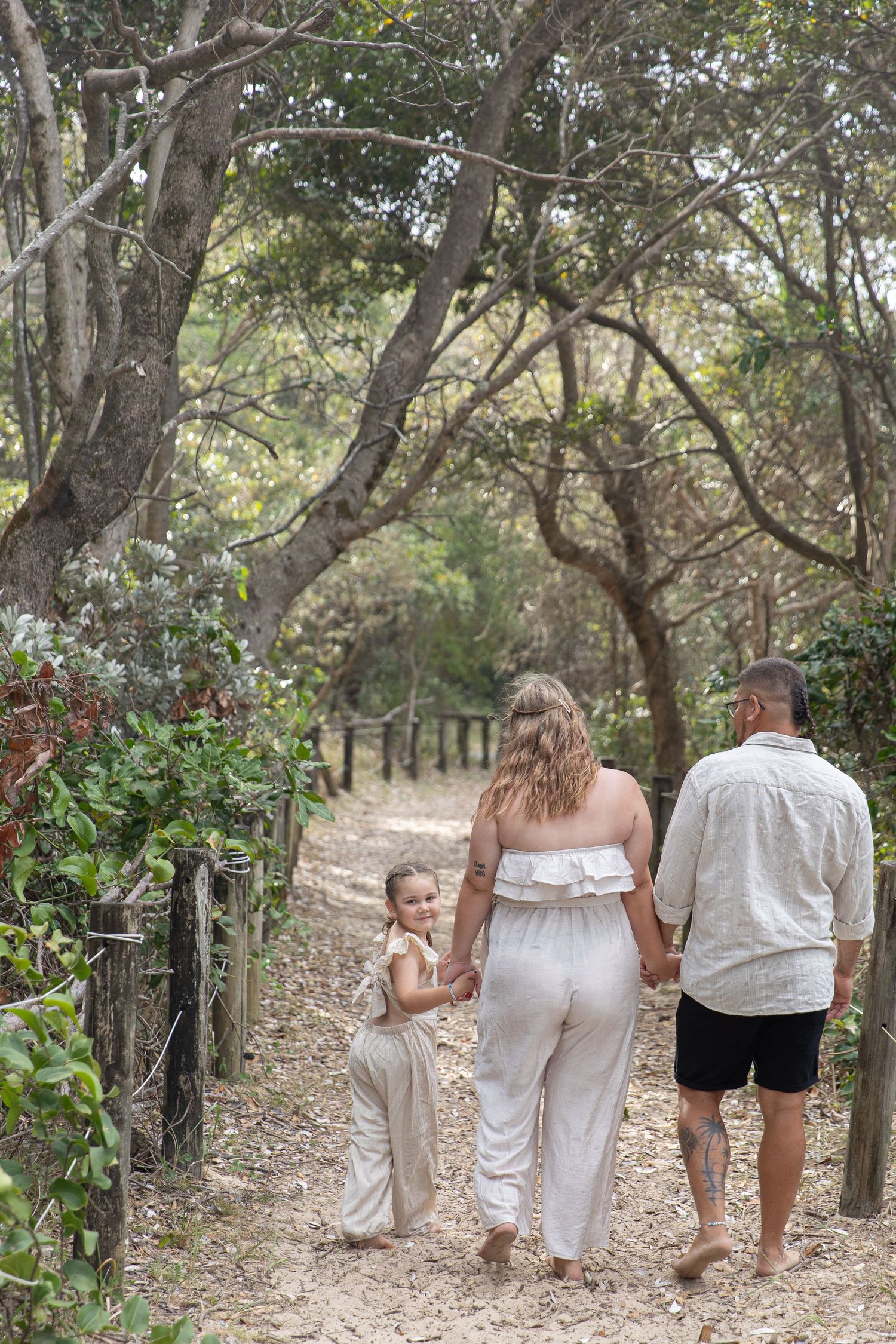 Family of three walking barefoot on a sandy forest path, young girl looking back, parents holding hands.