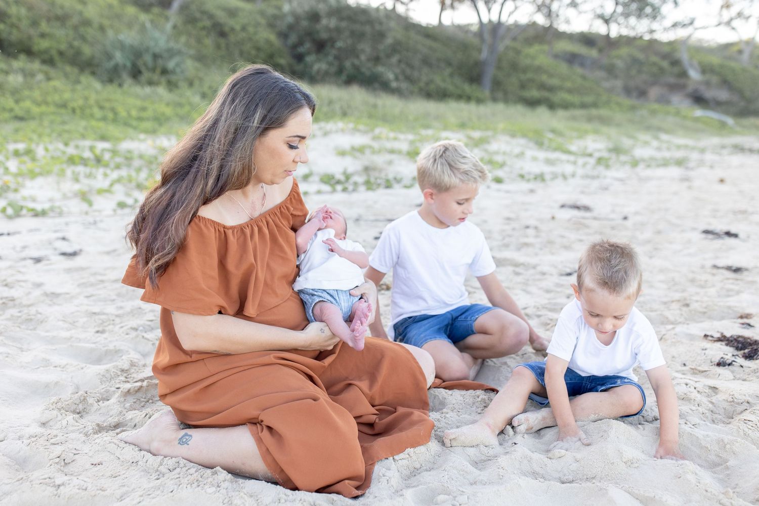 A mother in a rust dress sits on a sandy beach holding a newborn while two young boys play beside her.