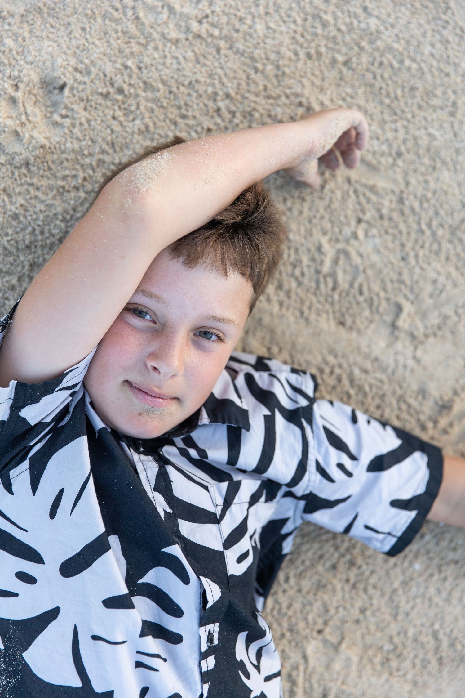 Child wearing black and white tropical print shirt lying on sandy beach.