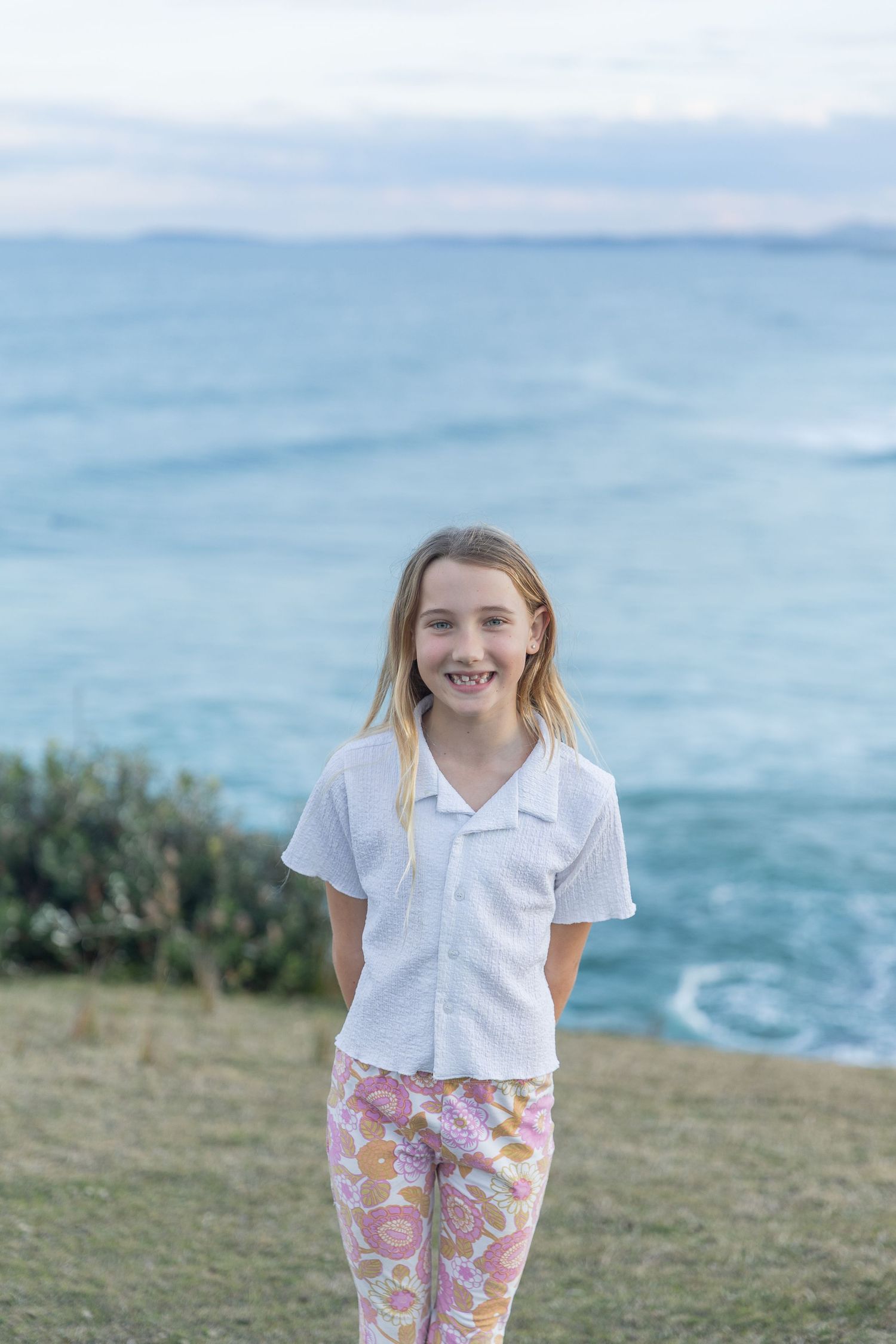 A person in a white shirt and floral pants stands on a grassy hill overlooking the ocean on a bright sunny day.