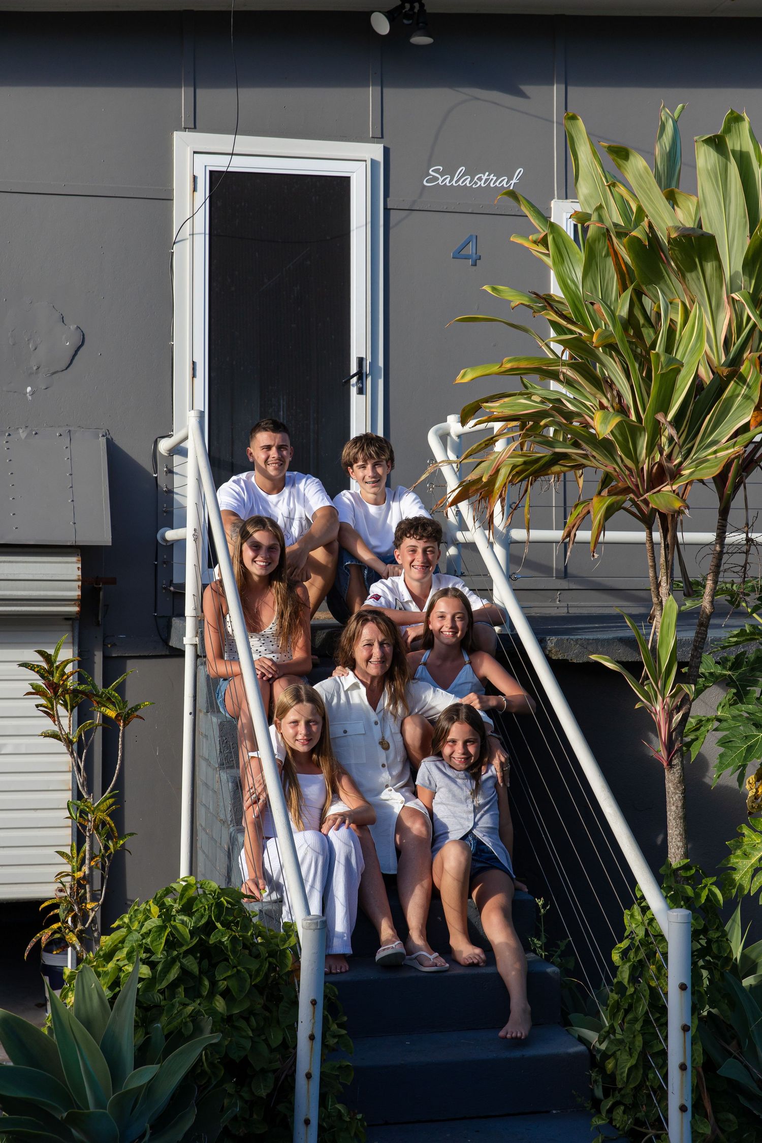 Large family in white outfits posing together on outdoor steps of a grey house surrounded by tropical plants.