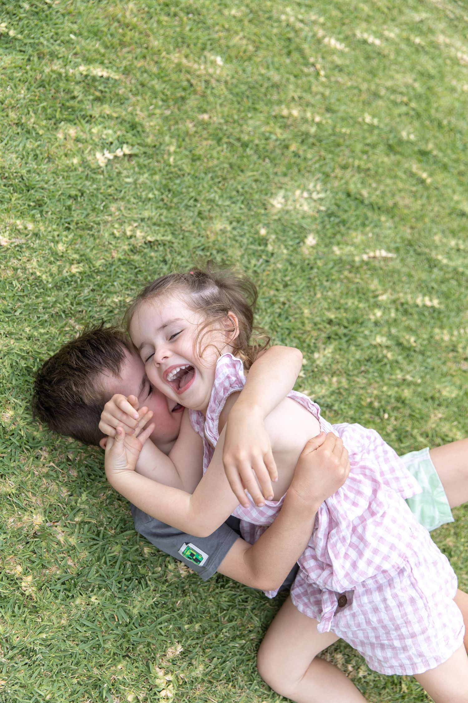 Playful siblings laughing and rolling on green grass during a fun outdoor moment.