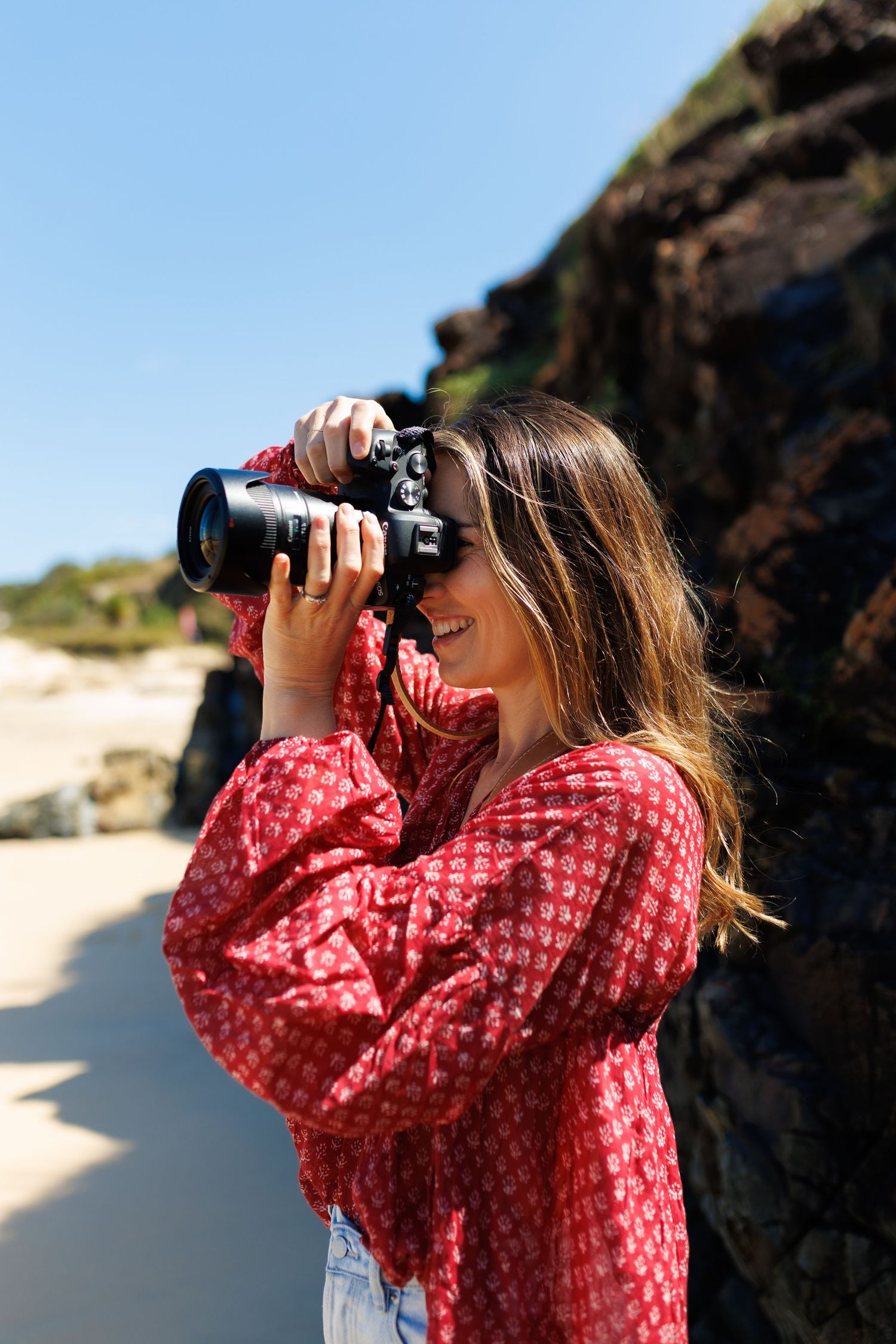 Person in red polka dot blouse using binoculars on beach with rocky cliffs.