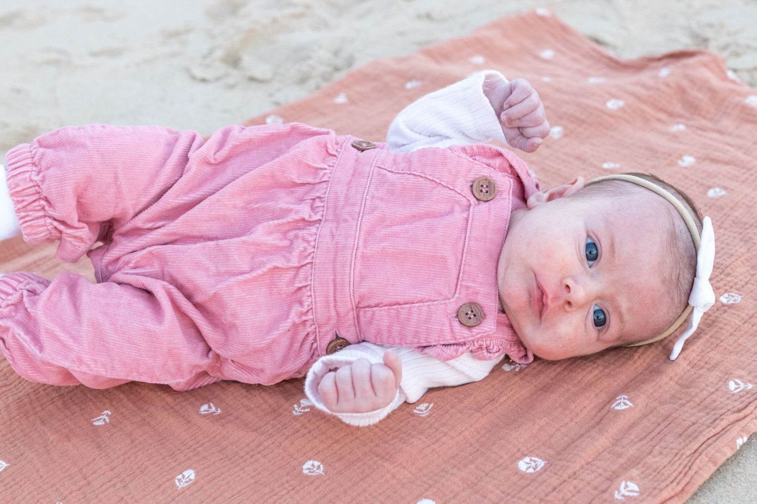 Adorable baby girl in pink corduroy overalls lying on a floral blanket outdoors with blue eyes.
