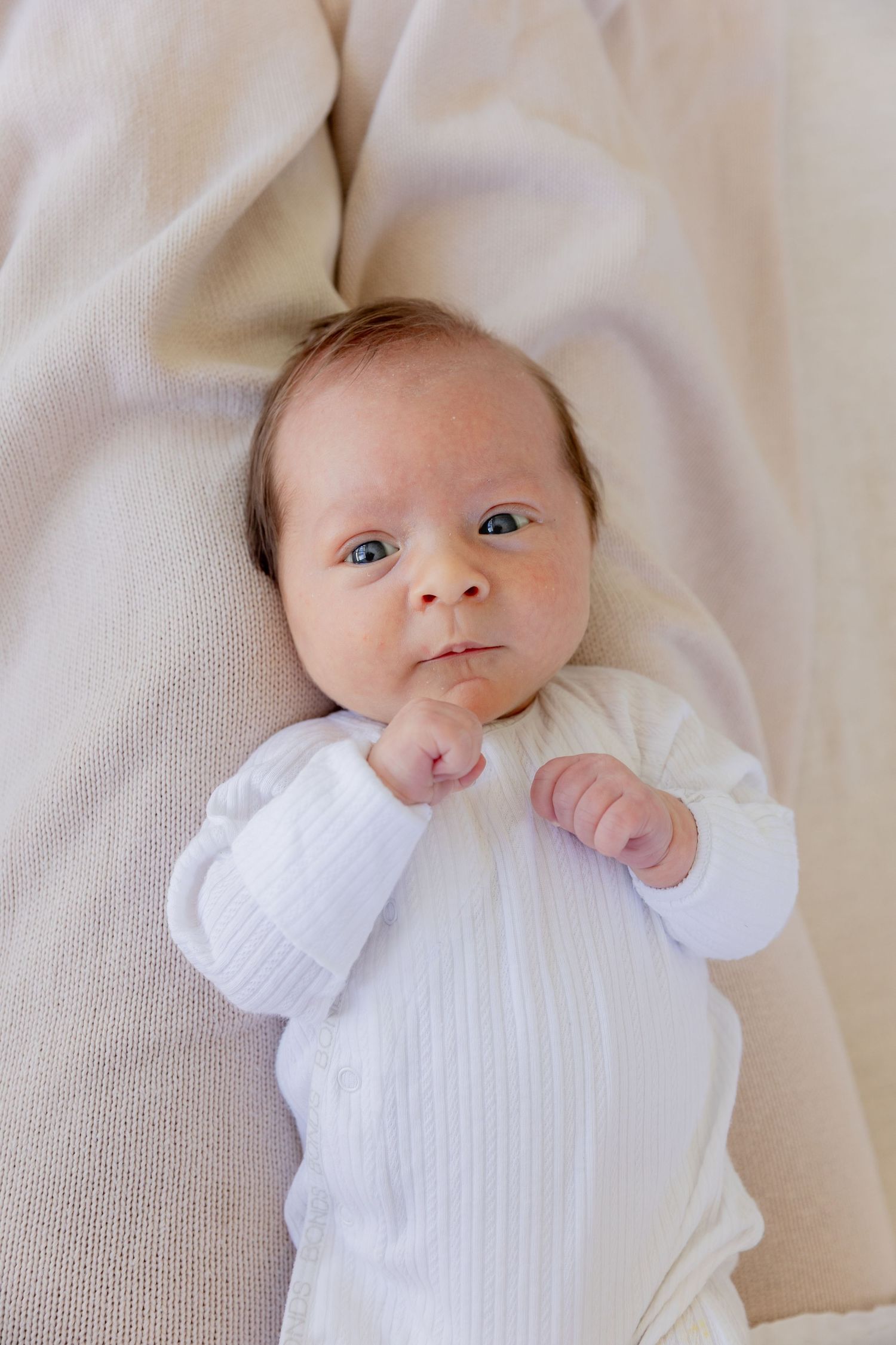 Baby wearing white outfit lies on beige bedding while sucking thumb.