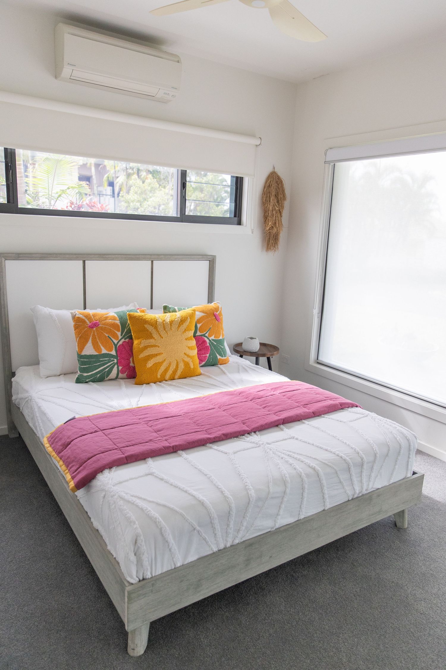 Modern bedroom with white bedding, colorful tropical pillows, and high windows.
