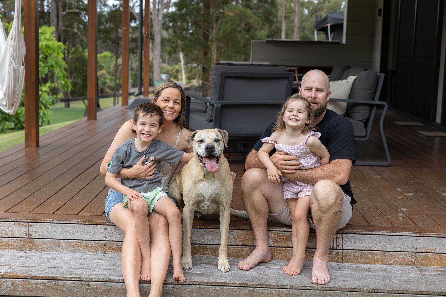 Family of four with a dog sitting on a wooden deck outside their home surrounded by lush greenery.
