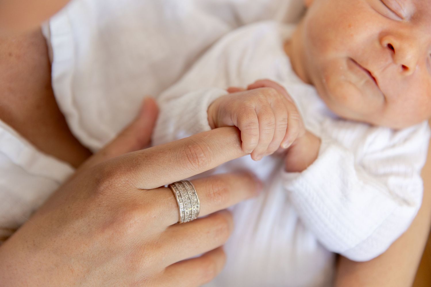 Newborn baby grasping mother's finger wearing a diamond wedding band, close-up tender moment.