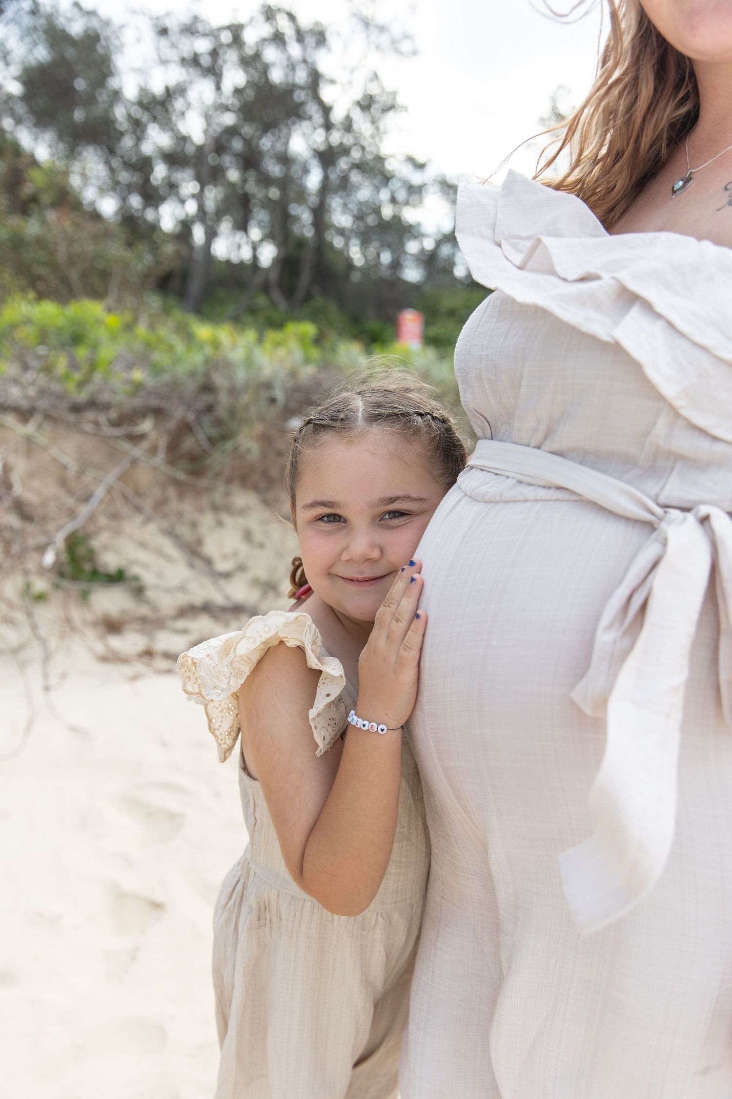 A child in a white dress stands next to someone wearing a white dress with a bow on a sandy beach.