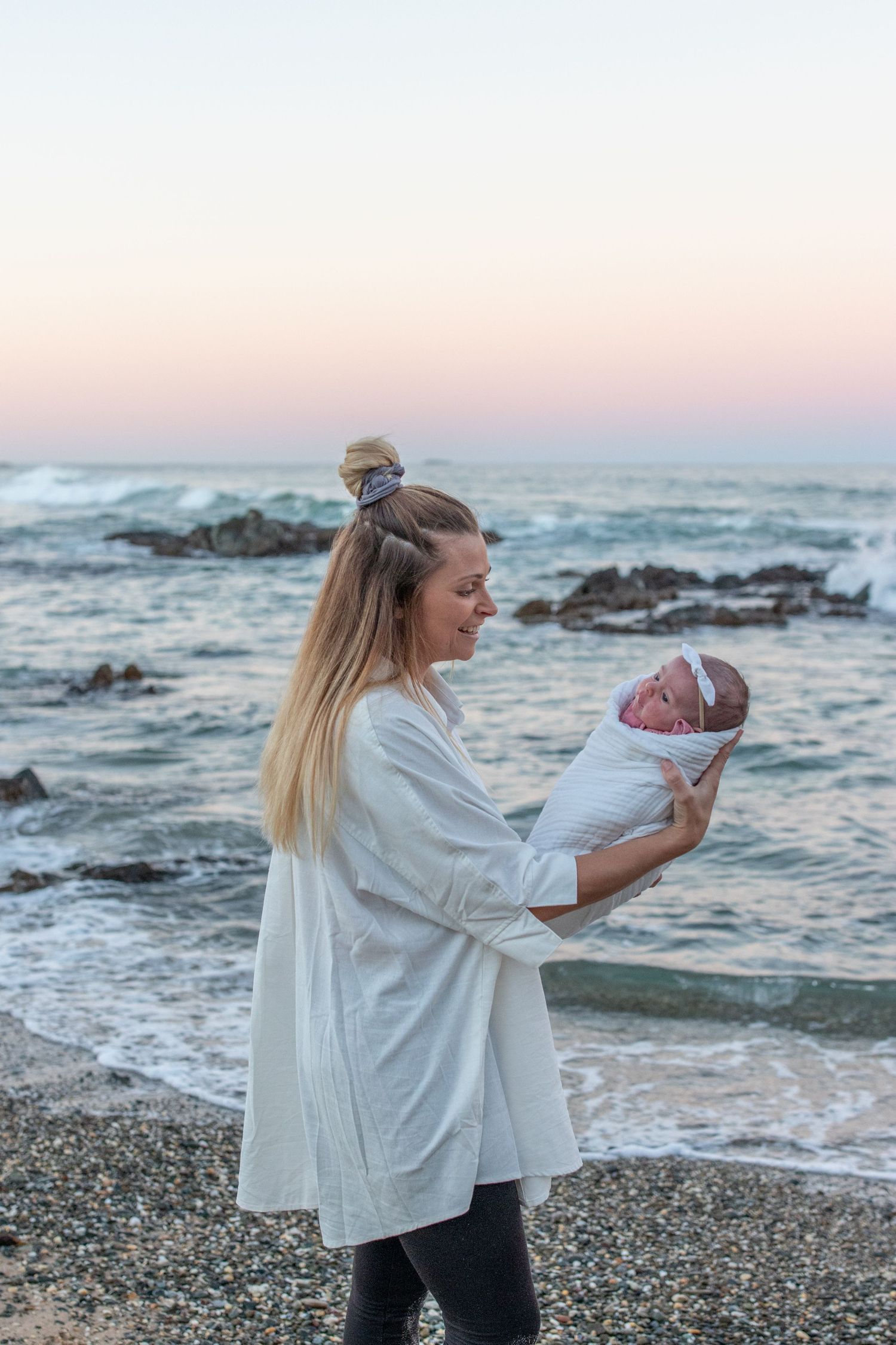 A mother holds her swaddled newborn baby on a rocky beach at sunset with ocean waves in the background.