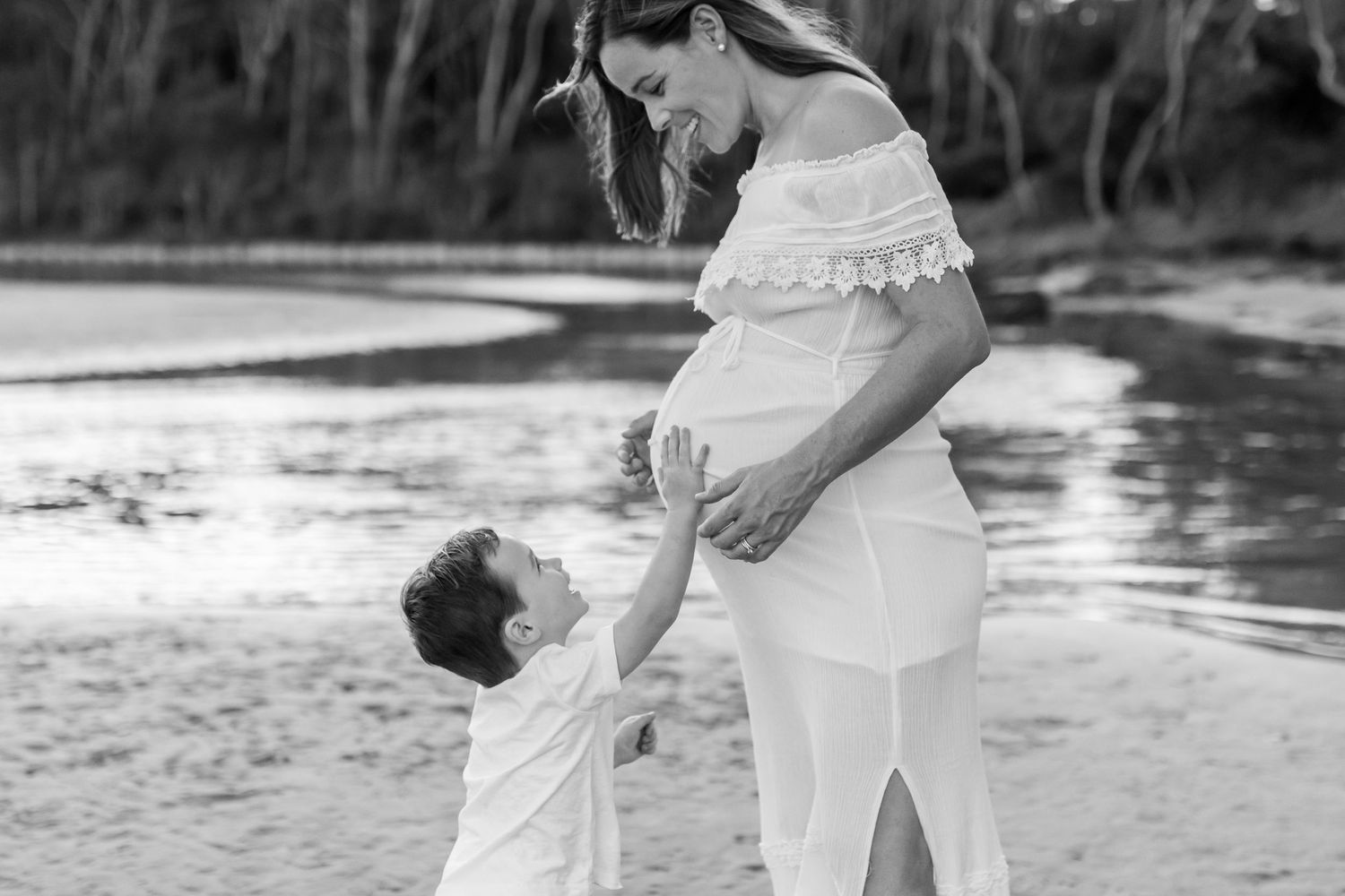 Black and white photo of pregnant woman in white dress with young child touching her belly near a river. Coffs Coast photographer.