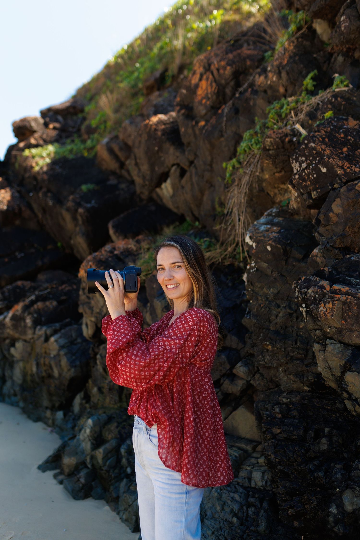 Woman in red polka dot blouse poses against rocky coastal backdrop on sandy beach.