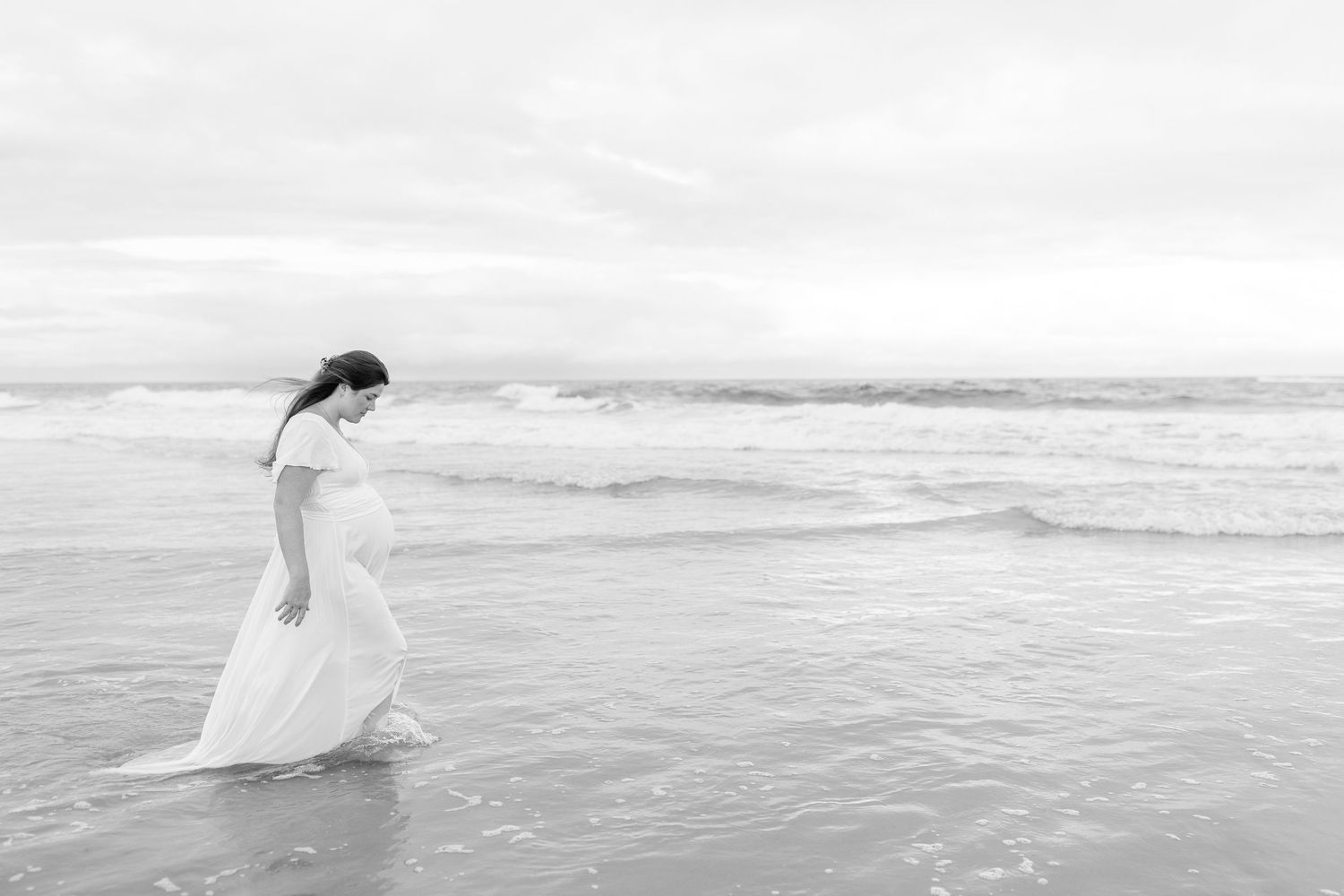 A pregnant woman in a white dress walks gracefully through ocean waves on a beach in a serene black and white photo.