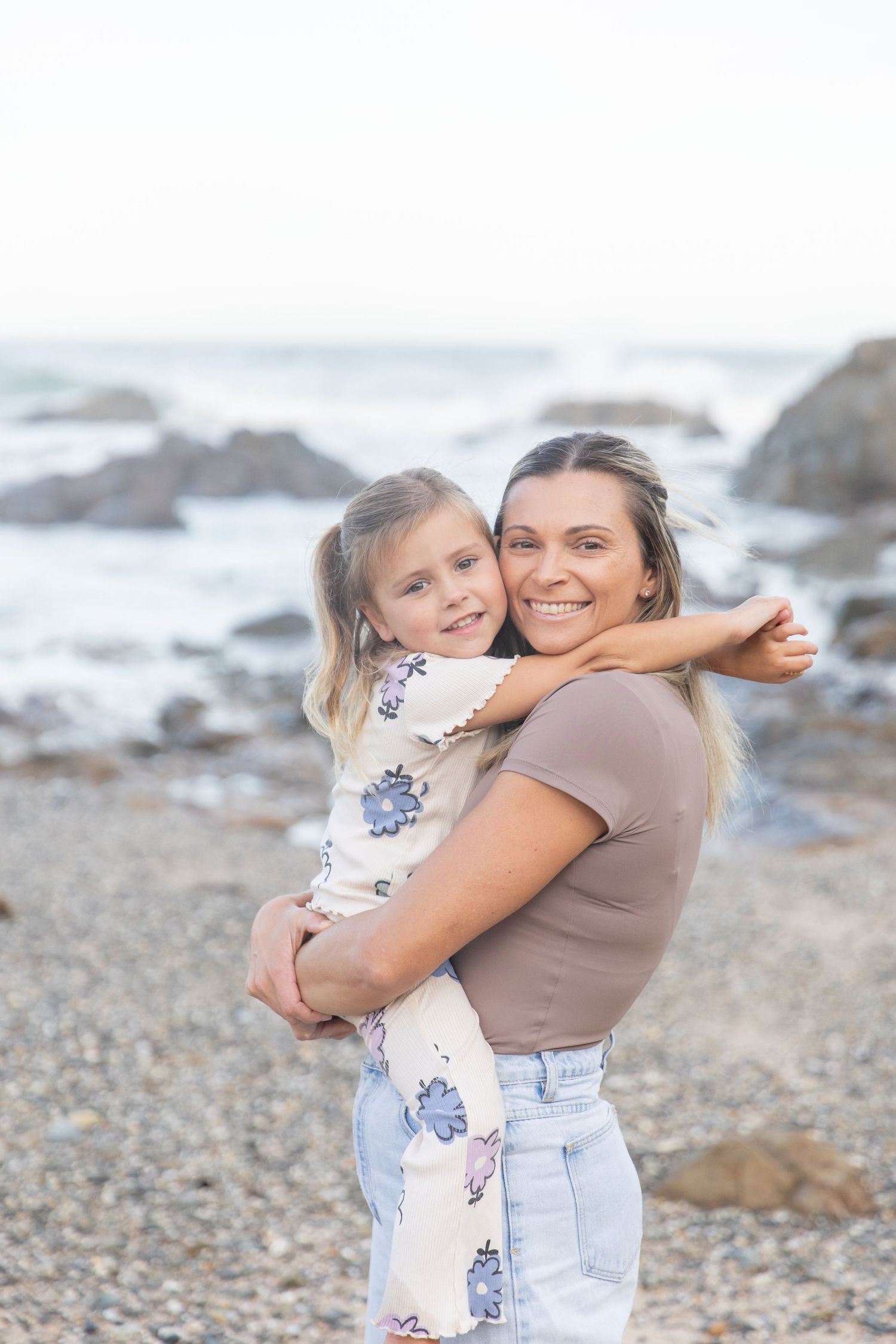 A smiling woman holds a young girl on a rocky beach with ocean waves in the background. Coffs Coast photographer.