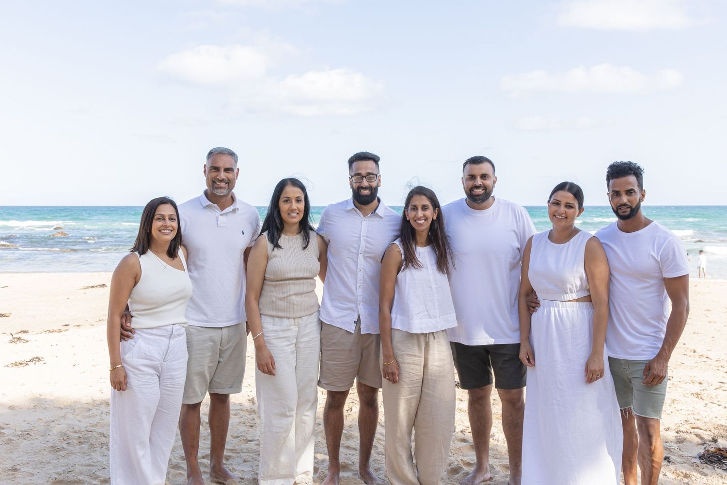 Family members posing together on a sunny beach, wearing white and neutral outfits with blue ocean in background. Coffs Coast photographer.