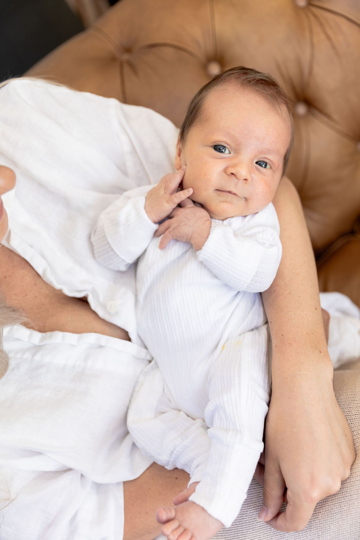 Baby in white clothing lies on tan leather couch during photoshoot.