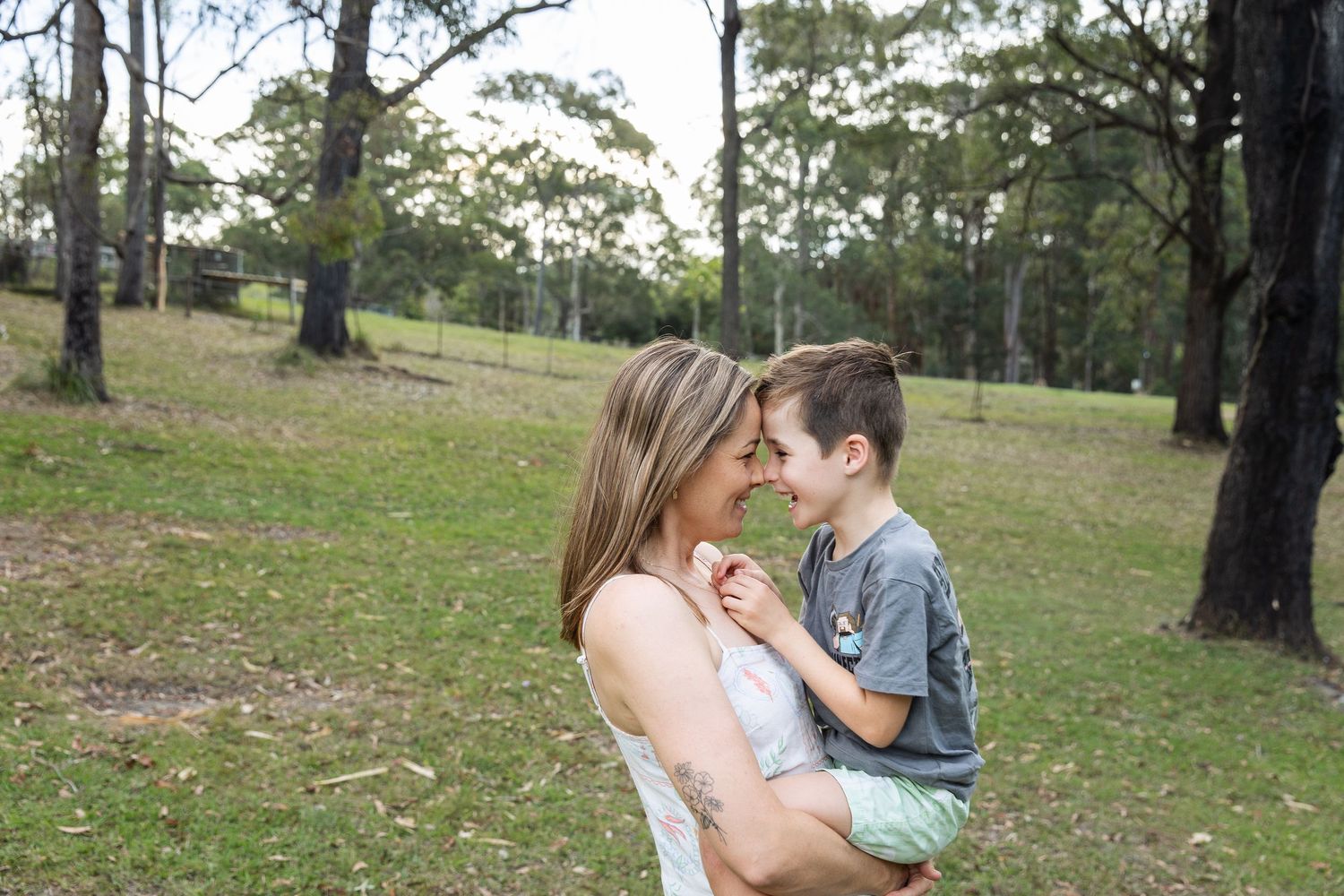 A mother holds her young son in a lush green park, sharing a tender nose-to-nose moment among tall trees.