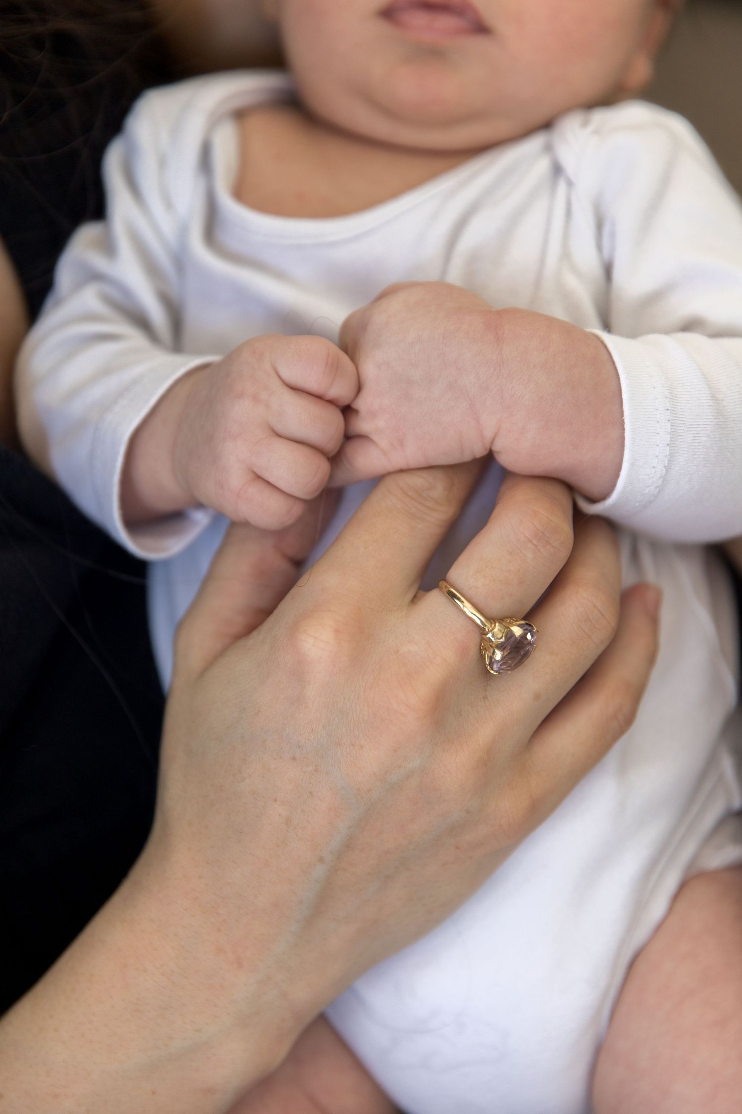 A mother's hand with a gold ring holds her baby in a white onesie, showcasing a tender parent-child bond.
