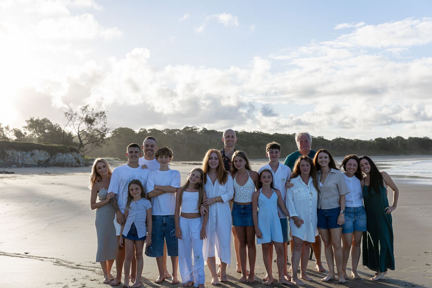 Large family group posing together on a sunny beach, wearing coordinated white and blue outfits, with ocean waves in the background.