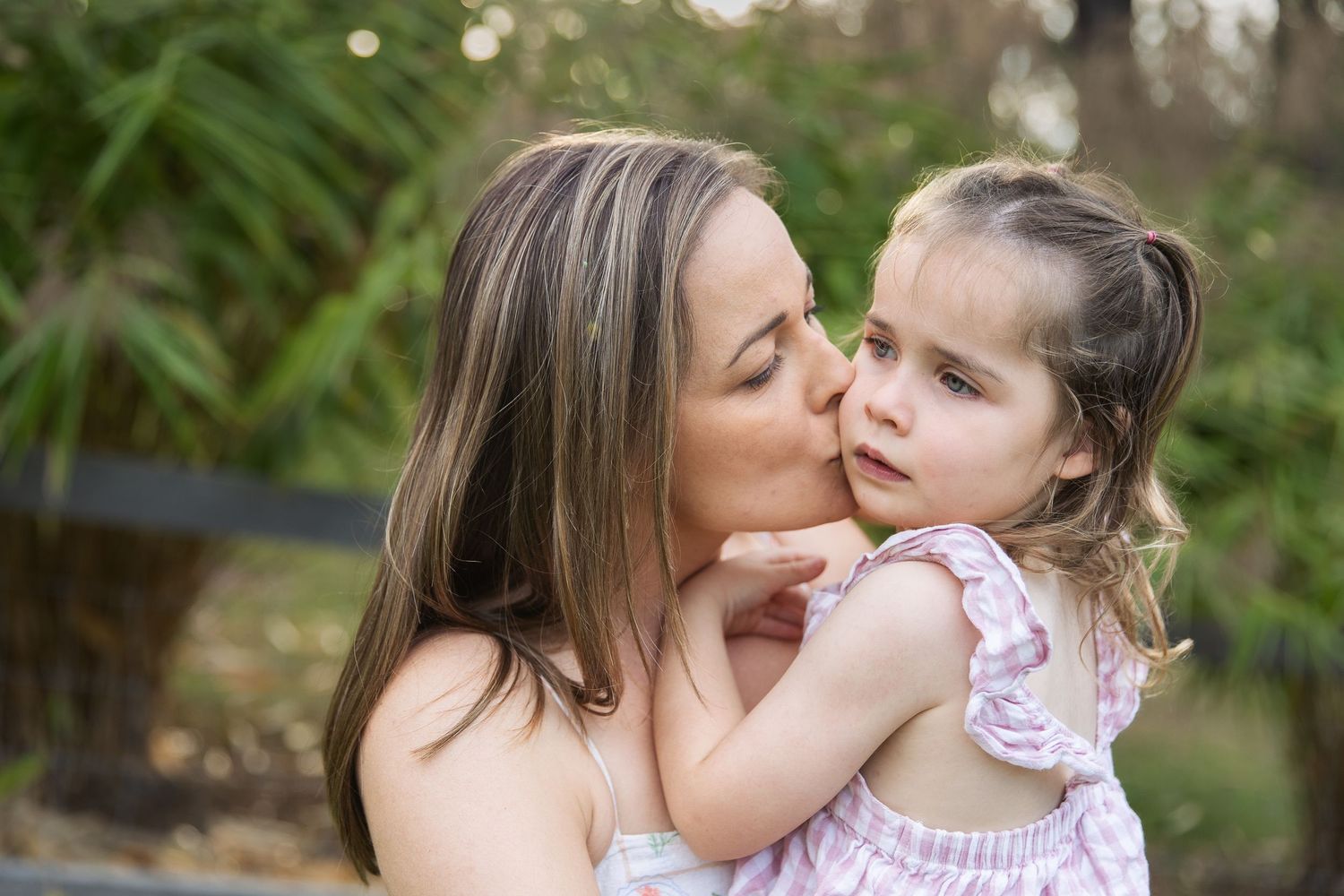 A mother tenderly kisses her young daughter on the cheek outdoors, both wearing light clothing.