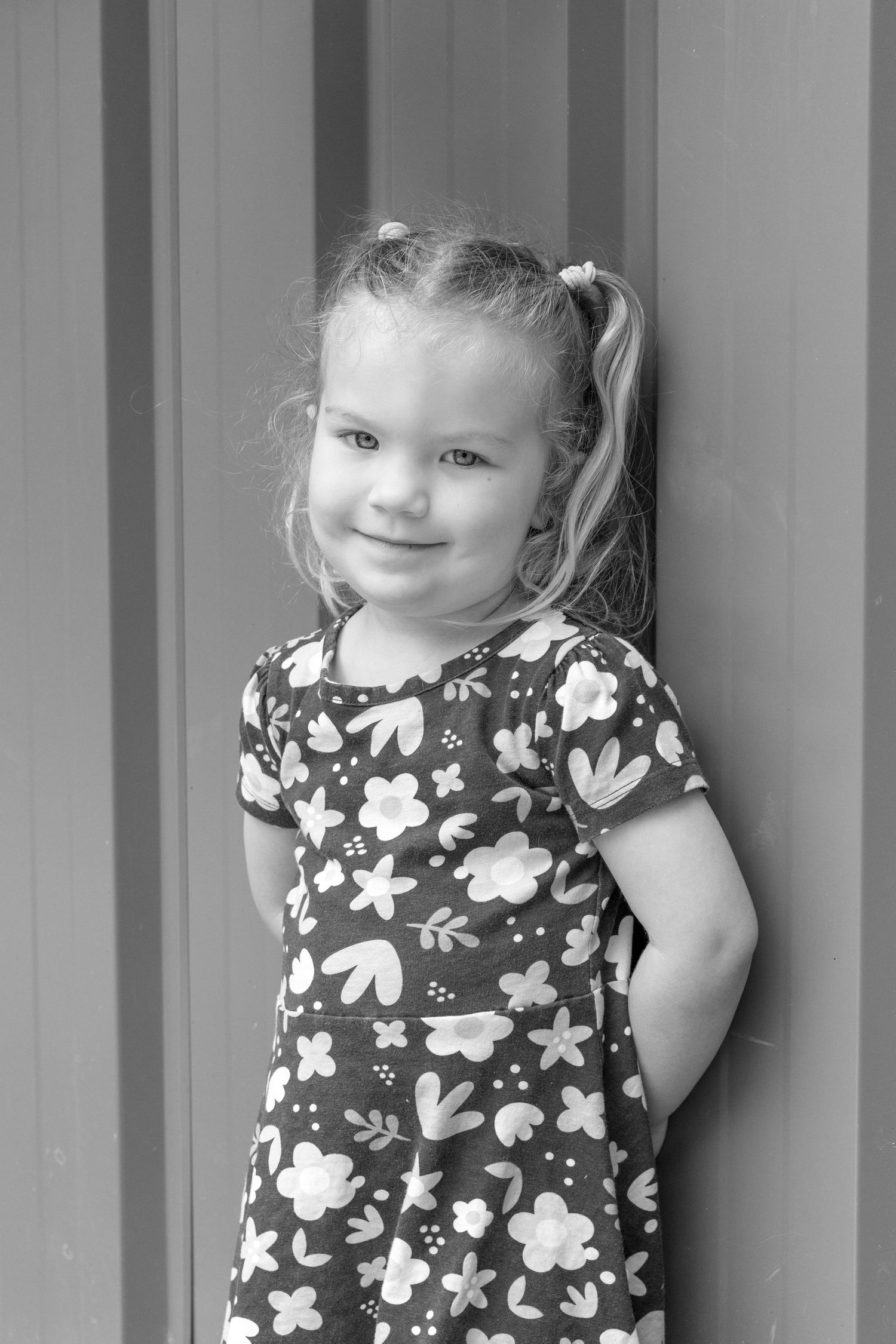Black and white portrait of a smiling young girl in a floral dress with pigtails, leaning against a striped wall. Coffs Coast photographer.