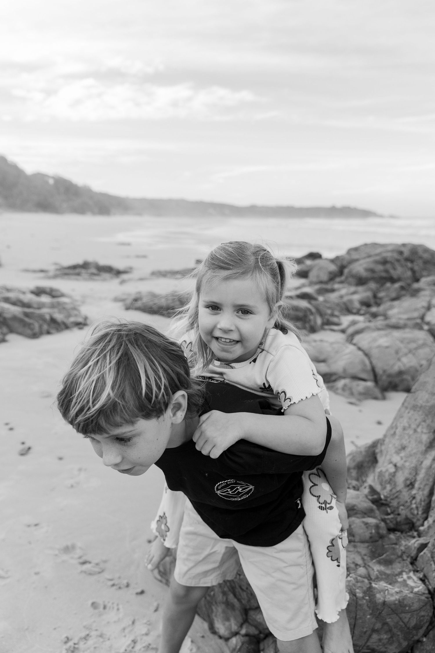 Black and white photo of a boy giving a smiling girl a piggyback ride on a rocky beach Coffs Coast photographer