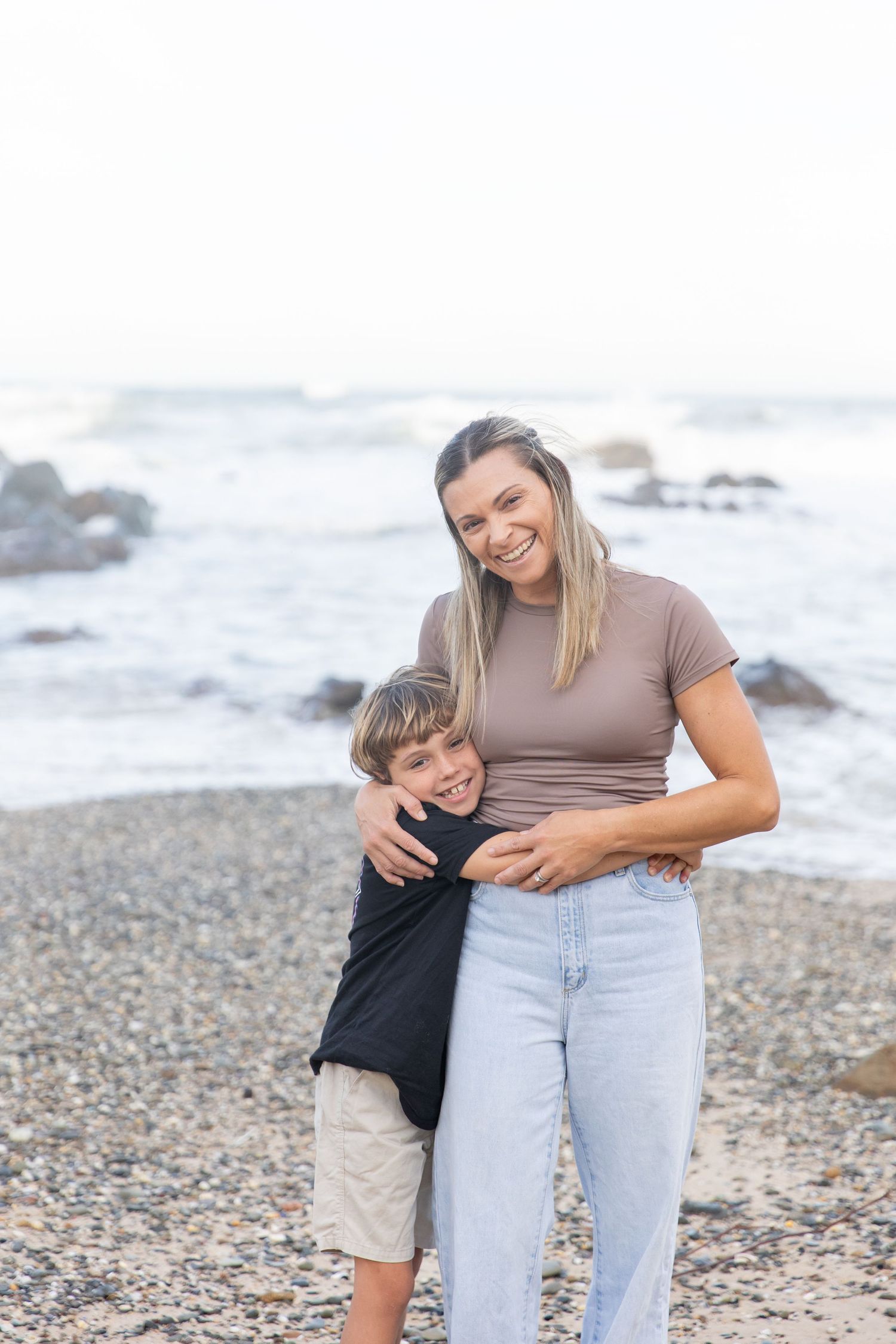 A smiling woman and young boy share a warm hug on a rocky beach with ocean waves in the background.