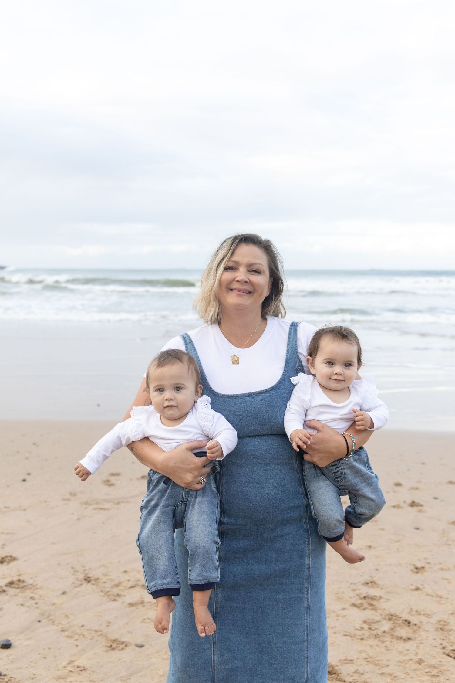 Woman in denim dress holding twin babies on sandy beach with ocean waves in background. Coffs Coast photographer.