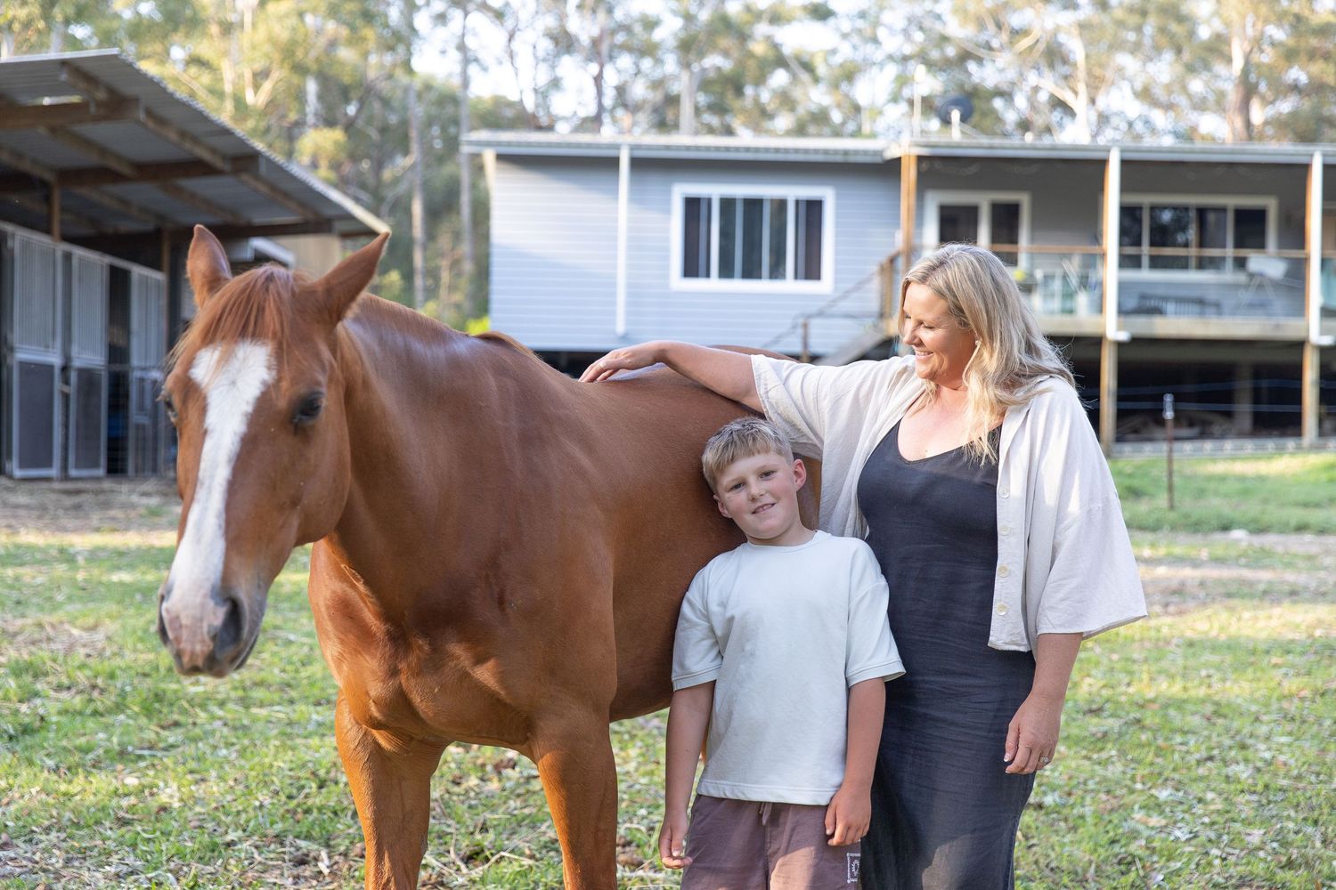 Family members standing near brown horse in sunny outdoor paddock setting.