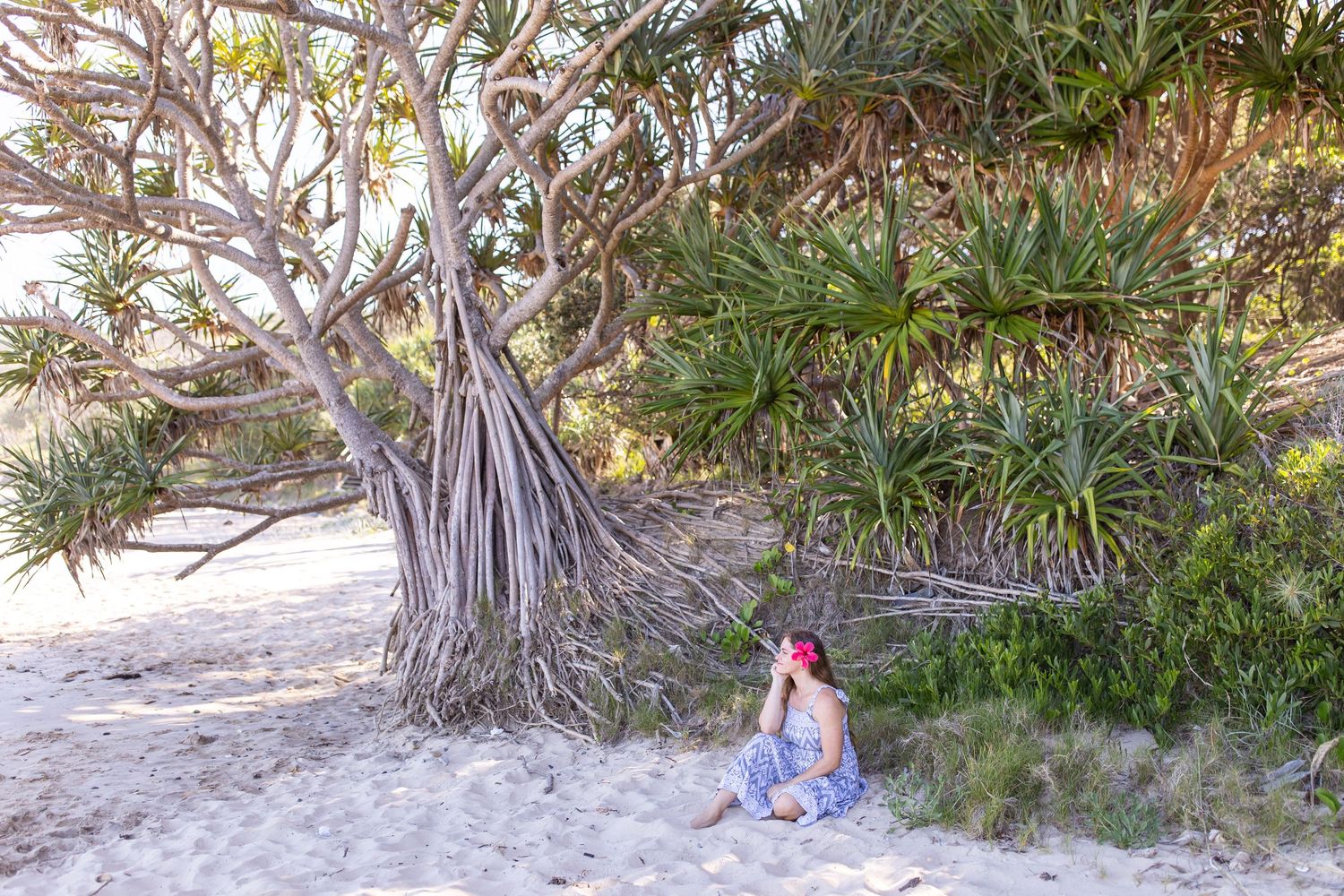 Woman sitting on sandy beach beside a large pandanus tree surrounded by lush tropical greenery.