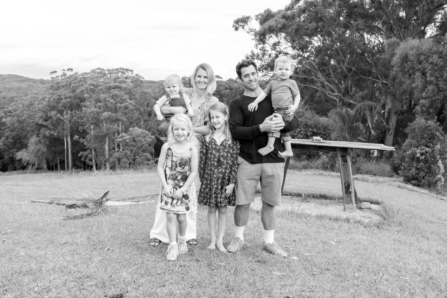 Black and white family portrait of parents with three young children outdoors on grass with eucalyptus trees. Coffs Coast photographer.