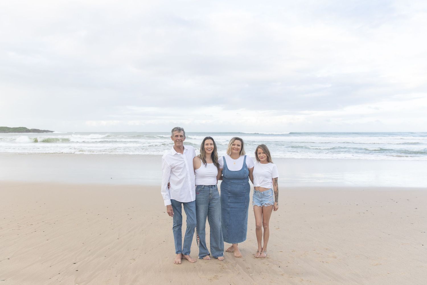 Family of four smiling together on a sandy beach with ocean waves in the background on a cloudy day.