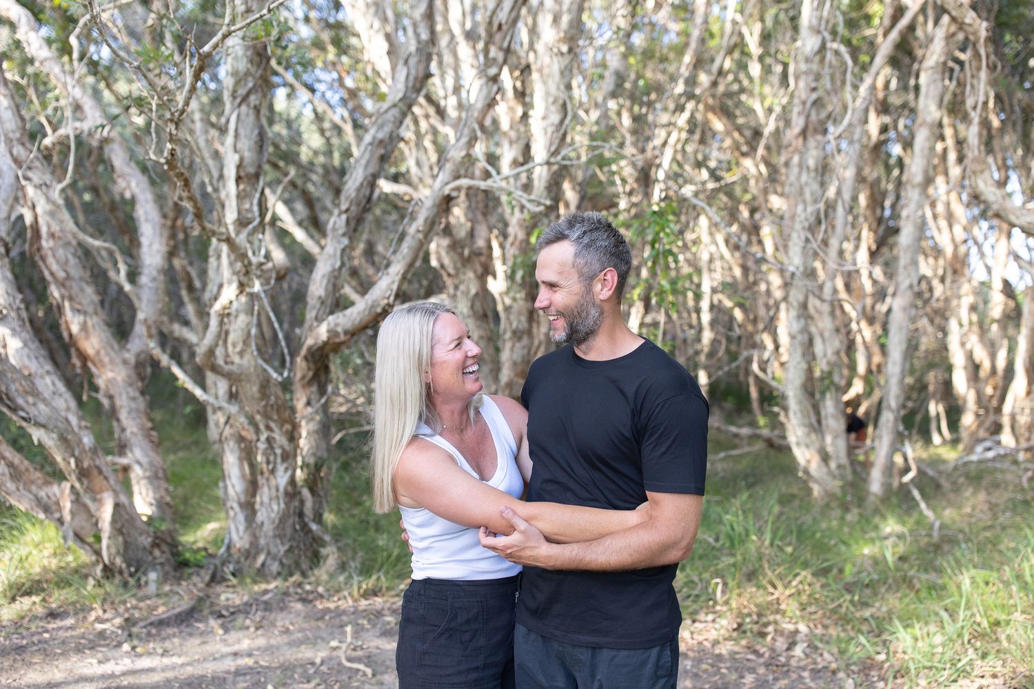 Couple smiling at each other while embracing on a forest path surrounded by bare white trees outdoors.