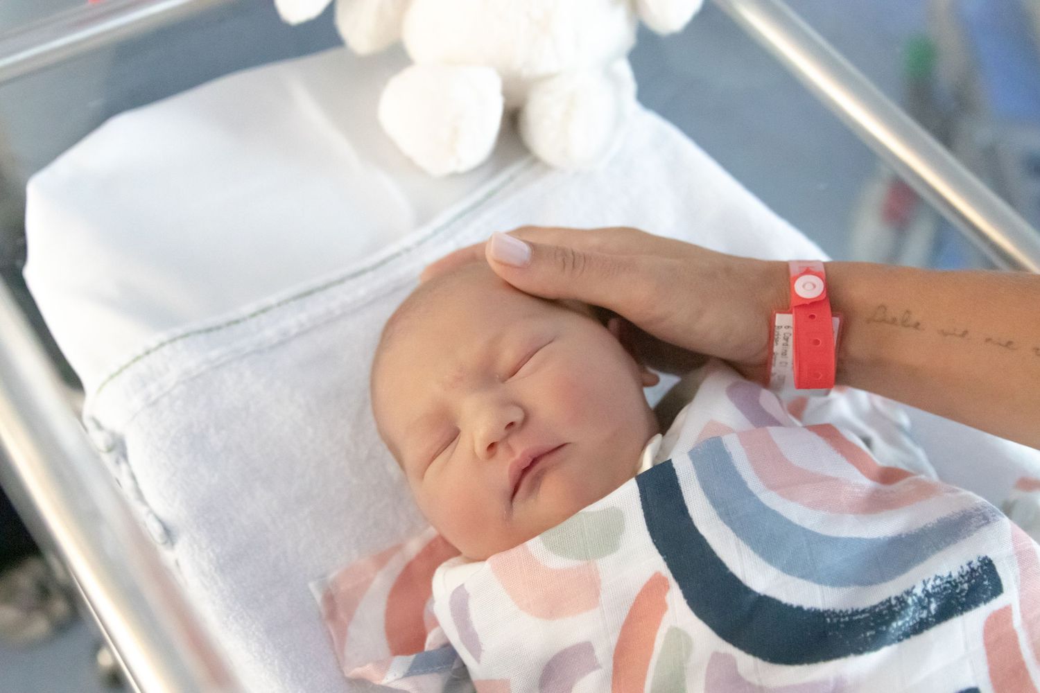 A newborn baby sleeping peacefully in a hospital bassinet while a parent gently touches their head.