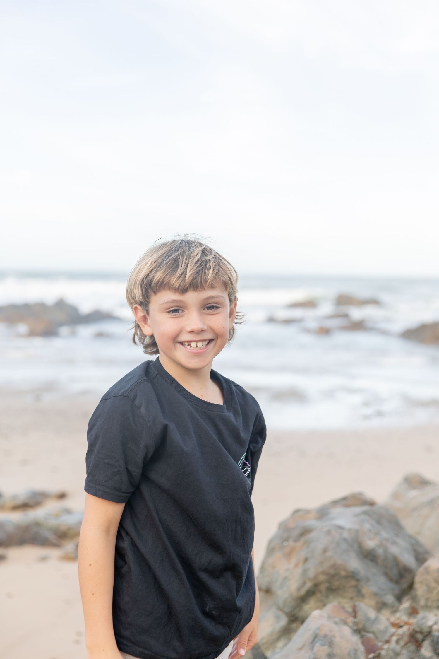 Smiling boy in black t-shirt standing on a rocky beach with ocean waves in the background Coffs Coast photographer
